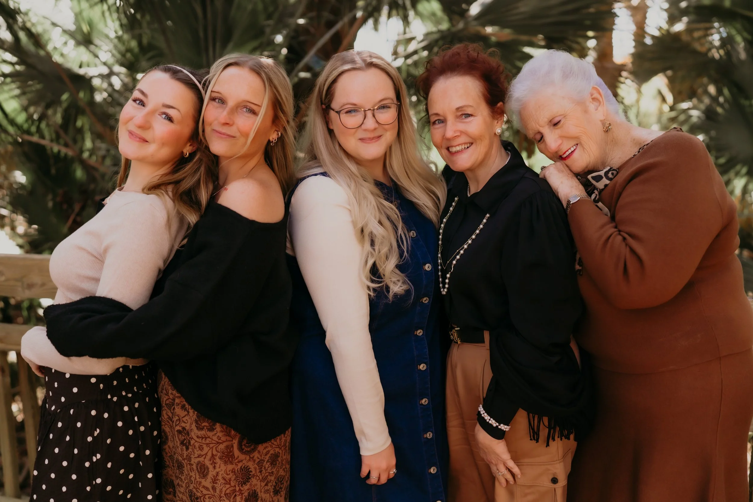 Six women standing close together outdoors, leafy background, smiling, embracing each other.