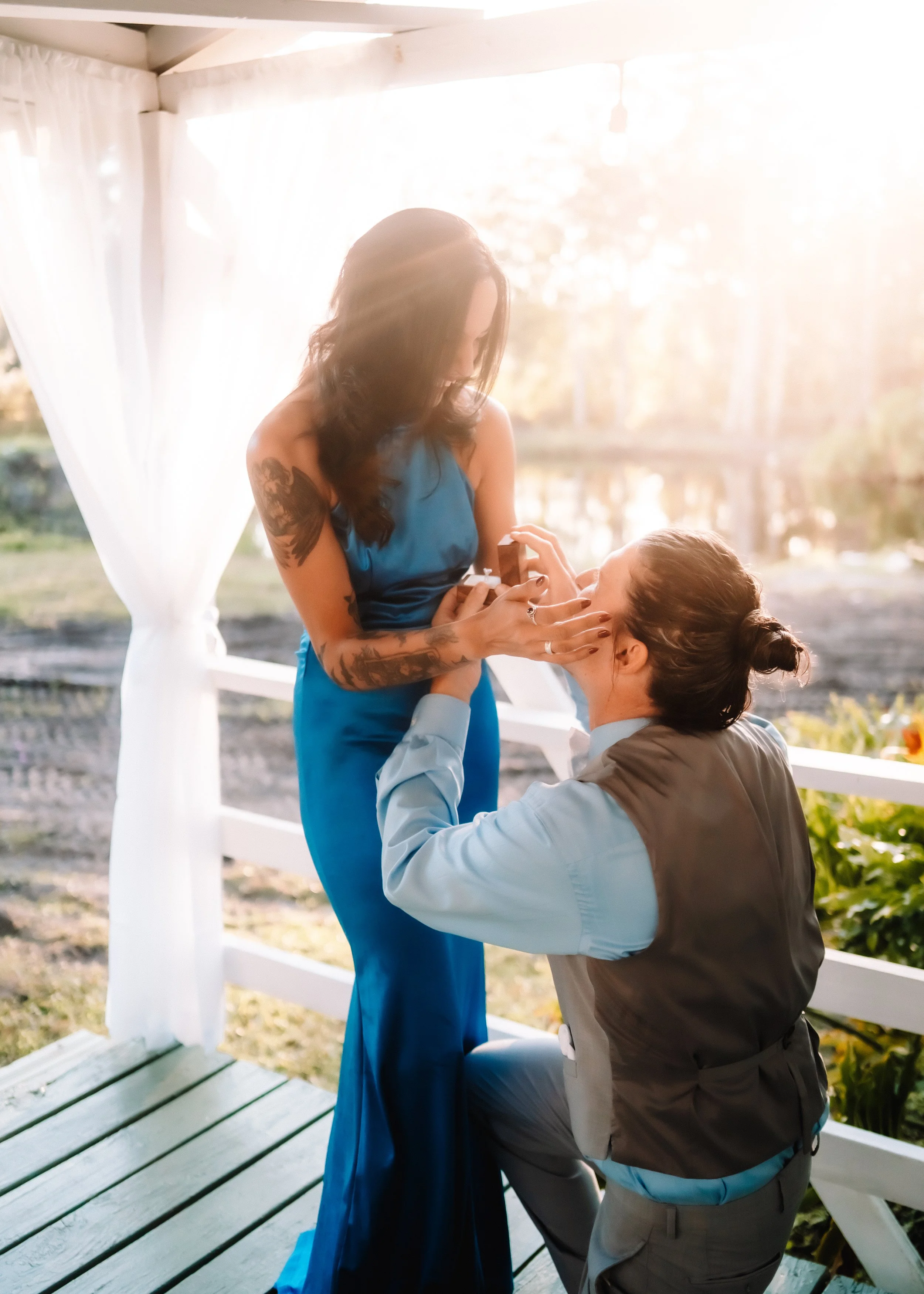 A woman with long dark hair and tattoos on her arms is smiling and looking at a man kneeling in front of her, holding her hands. The woman is wearing a blue dress, and the man is wearing a light blue shirt and gray vest. They are in a bright outdoor 