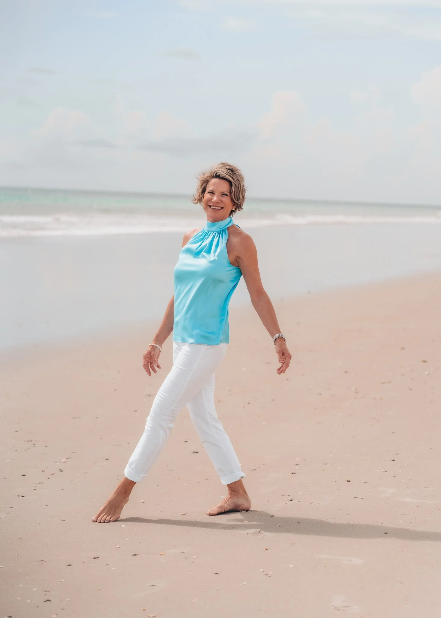 A woman walking barefoot on a sandy beach, smiling at the camera, with ocean waves and a cloudy sky in the background.