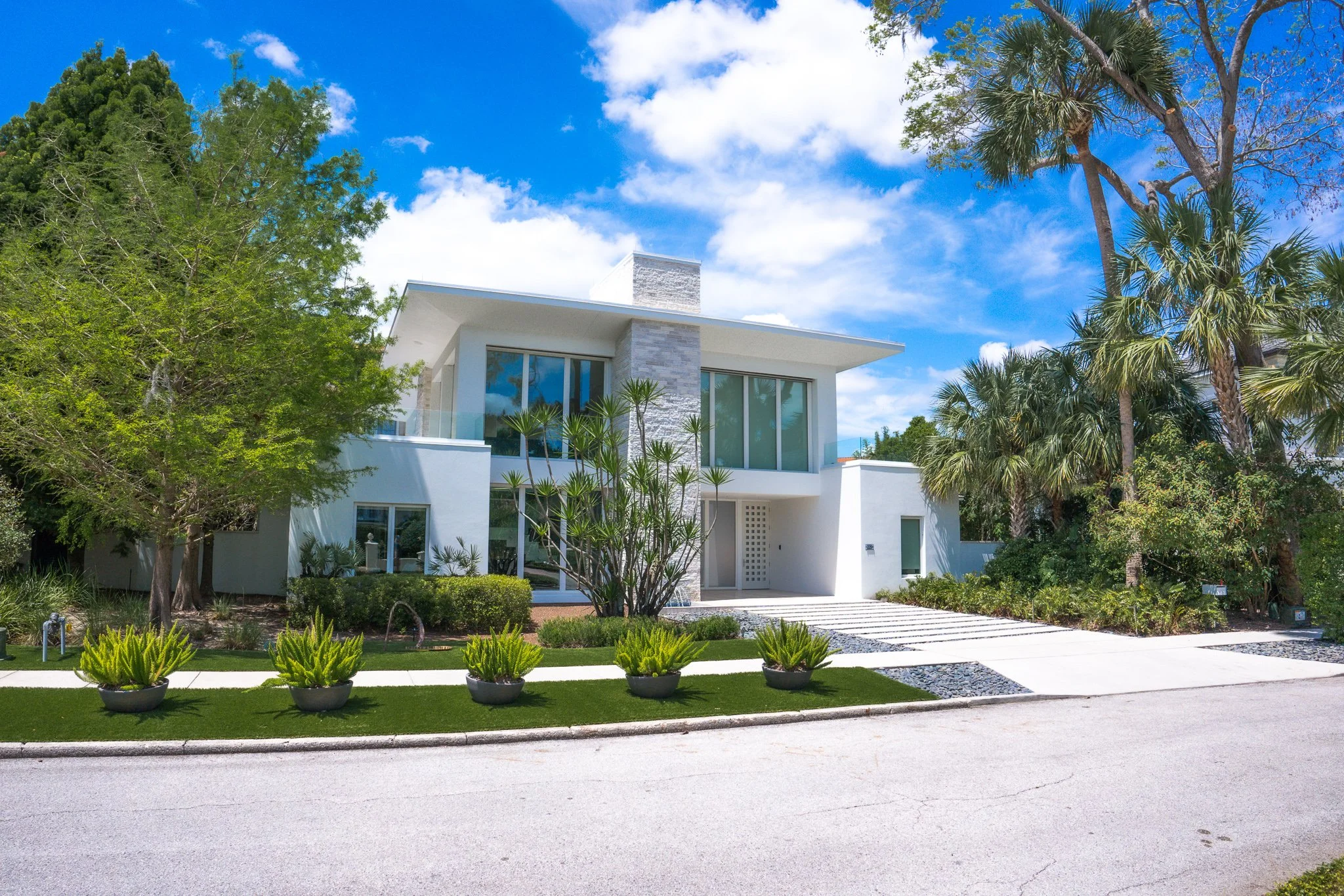 Modern white house with large glass windows, surrounded by greenery and palm trees, under a partly cloudy sky.