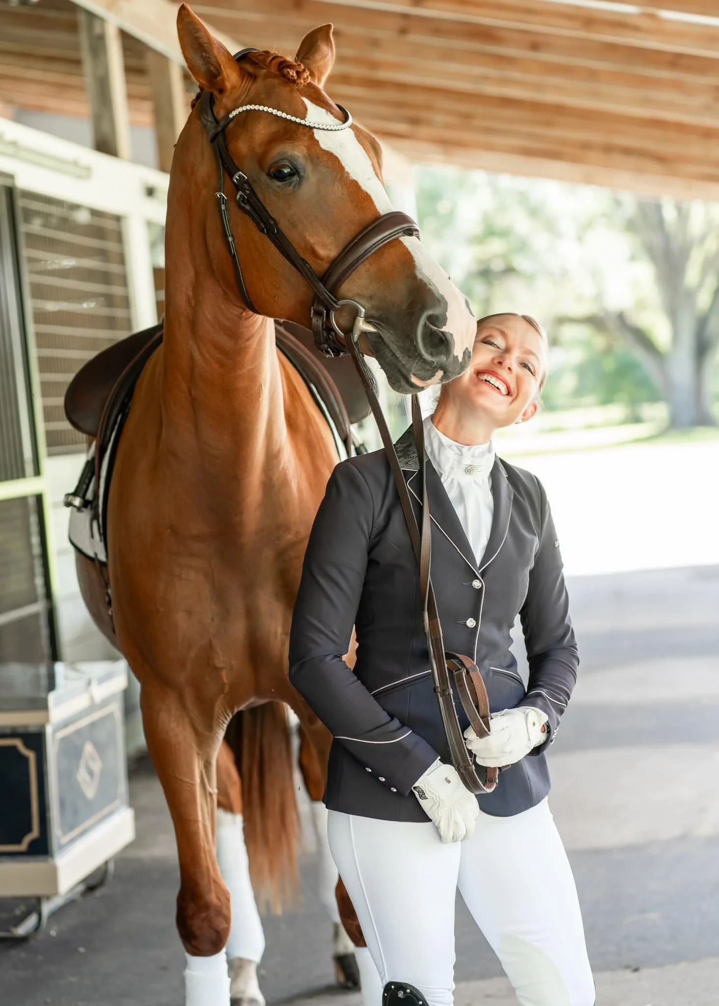 A woman in equestrian riding attire standing next to a chestnut horse, smiling with her head tilted toward the horse, in a stable or riding arena.