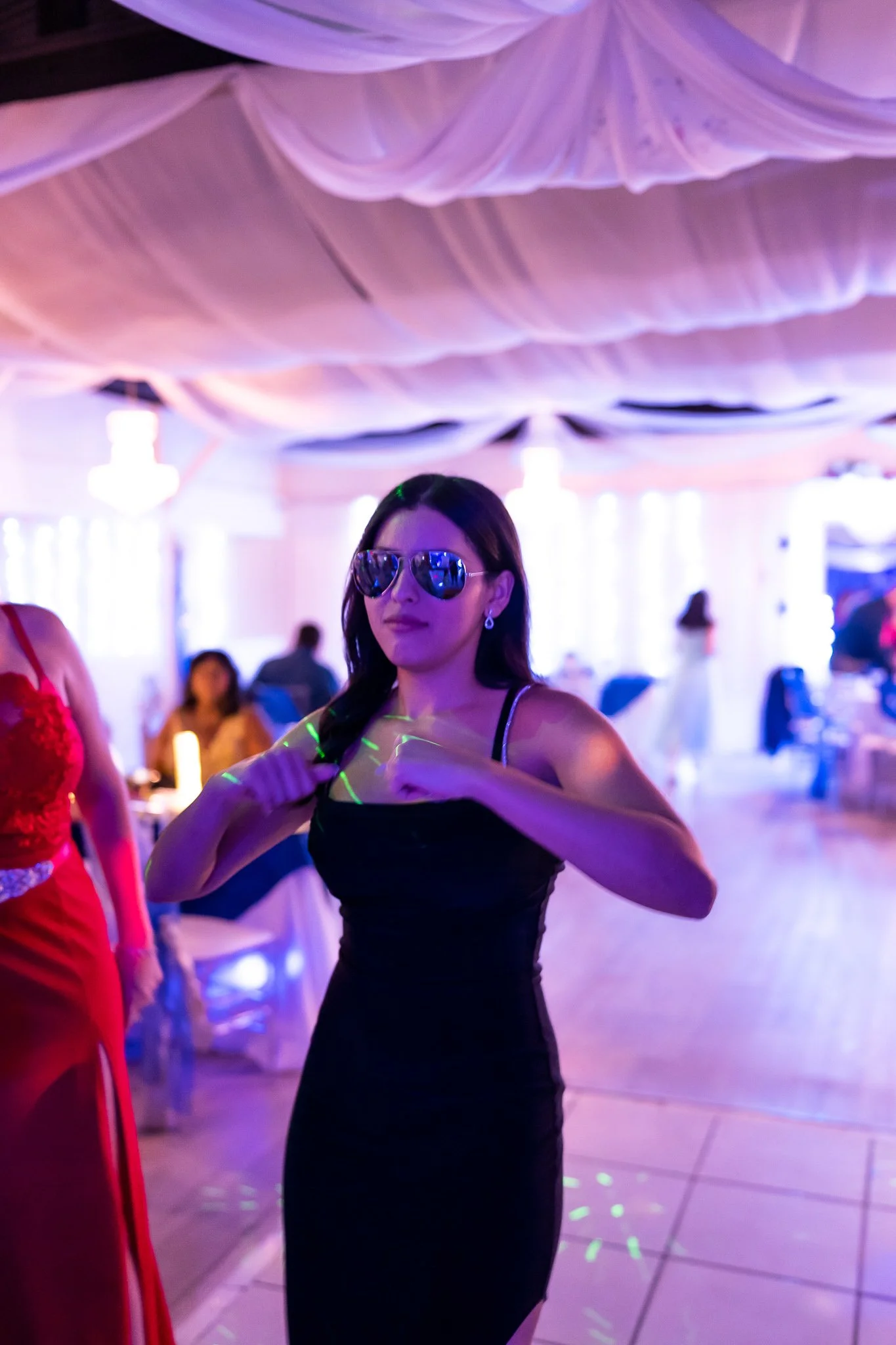 A woman with long dark hair wearing sunglasses and a black dress is at a party with purple and pink lighting, standing in a decorated indoor venue.