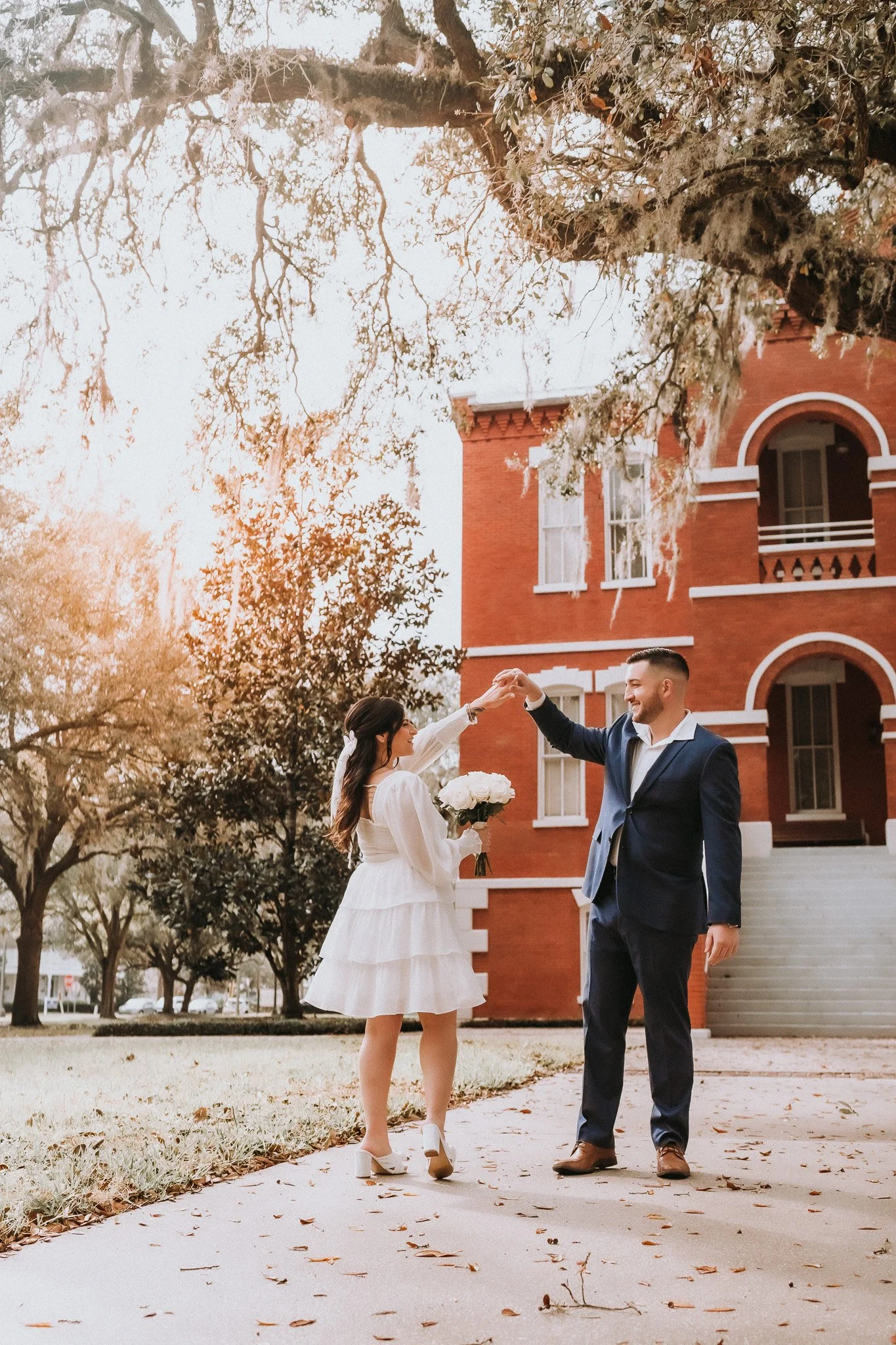 A bride and groom dancing outdoors in front of a red brick building with white trim. The bride is wearing a white lace dress and holding a bouquet of white flowers. The groom is wearing a navy blue suit and white shirt. They are smiling and holding h