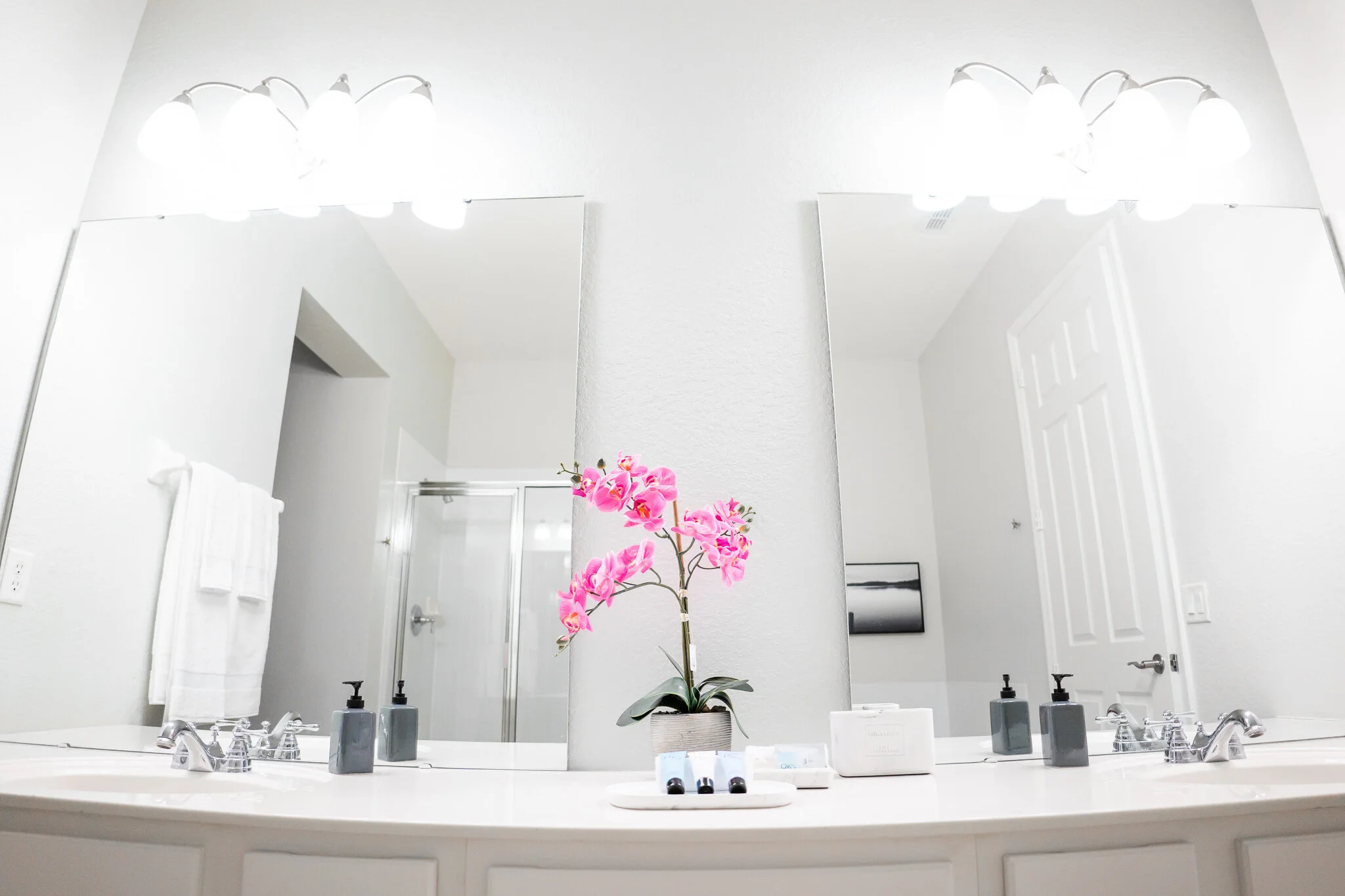 A bathroom vanity with a large mirror, two sinks, pink orchid flower in a pot, soap dispensers, toiletries, and a white countertop.