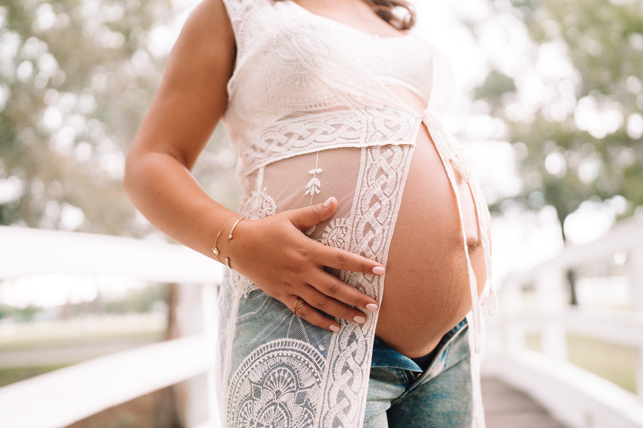 Close-up of a pregnant woman wearing a white lace top, with her hand resting on her belly, standing outdoors with blurred trees in the background.