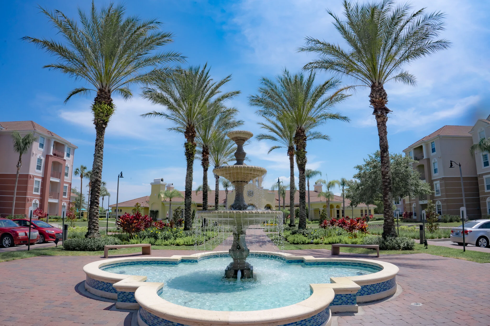 A picturesque courtyard with a water fountain in the center, surrounded by tall palm trees and landscaped gardens, with residential buildings and parked cars in the background under a clear blue sky.