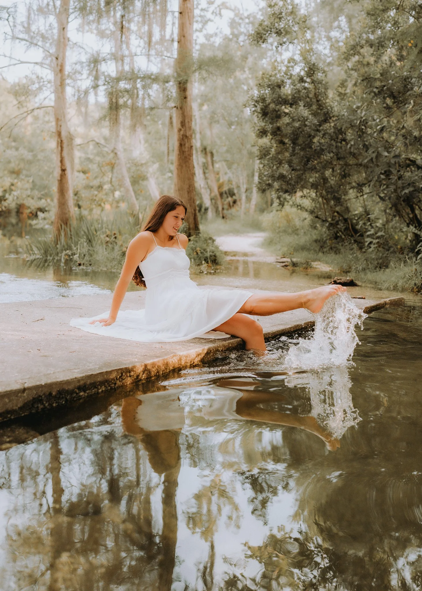 A young woman in a white dress sitting on a concrete ledge by a creek, splashing water with her foot, surrounded by trees and greenery.