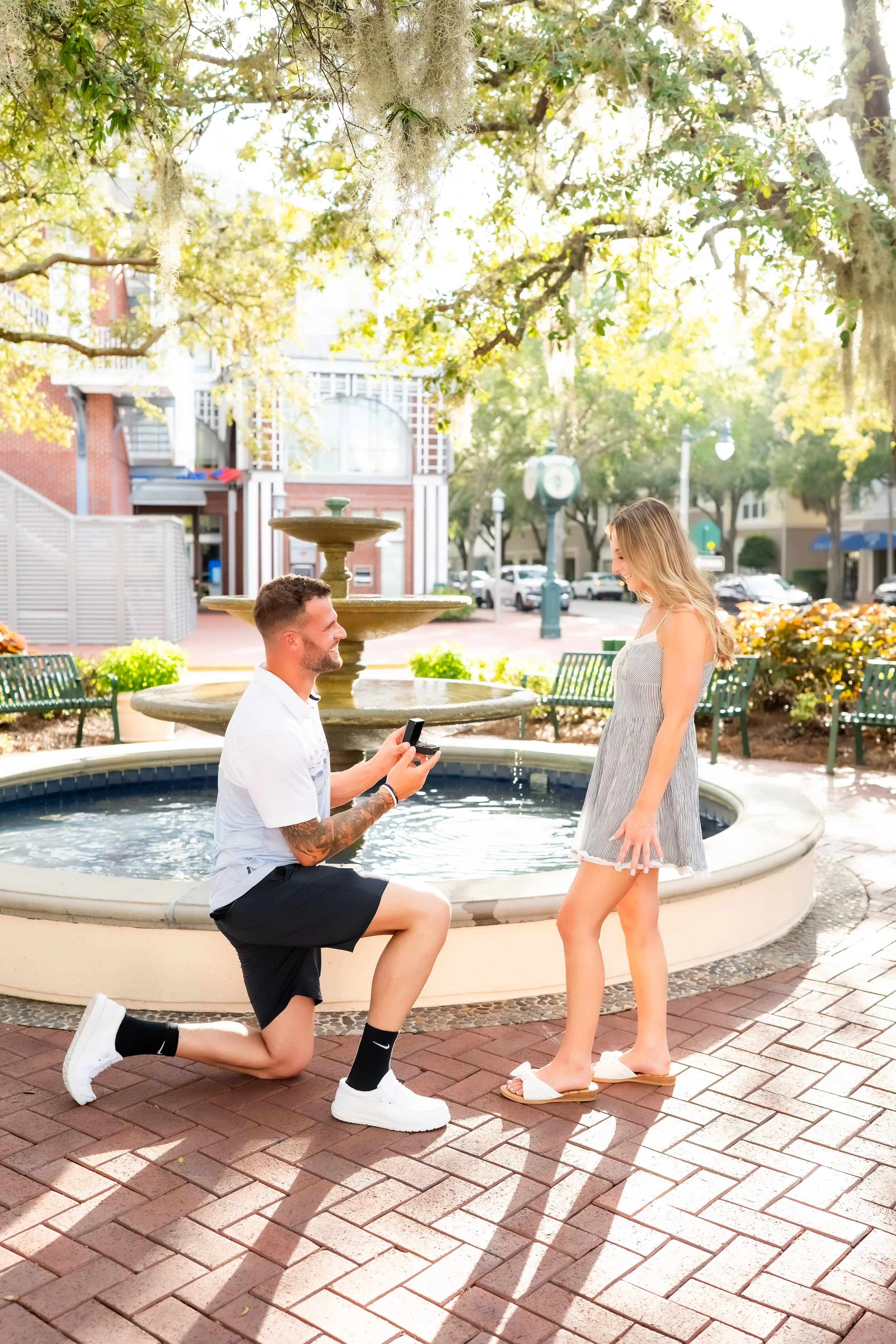 A man kneeling on one knee in front of a woman, proposing marriage by holding a ring box. The woman is smiling, standing in front of a fountain in an outdoor urban park setting during daylight. The scene is bright with sunlight filtering through tree