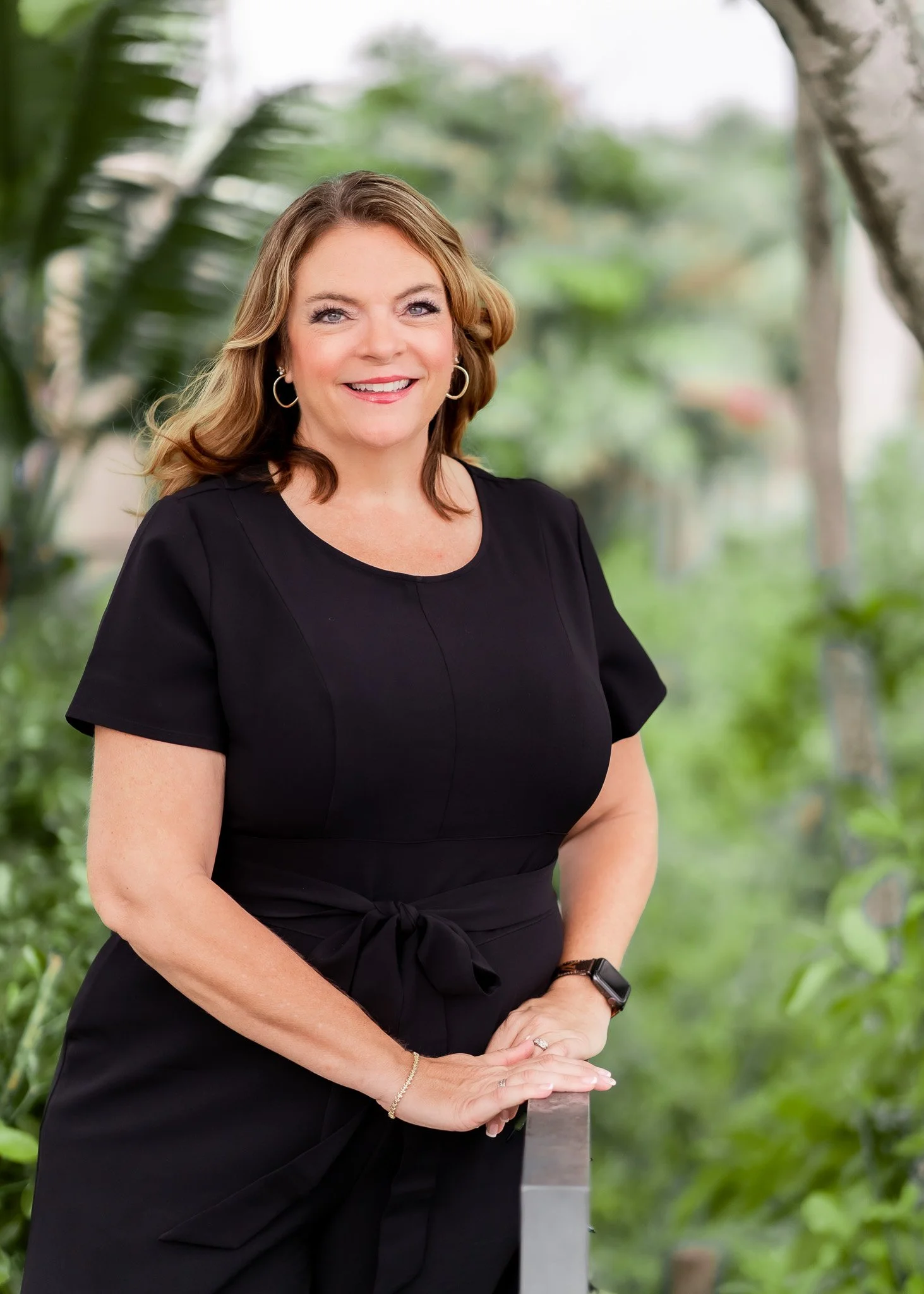 A woman with shoulder-length light brown hair, wearing a black dress, standing outdoors with greenery in the background, smiling and leaning on a railing.