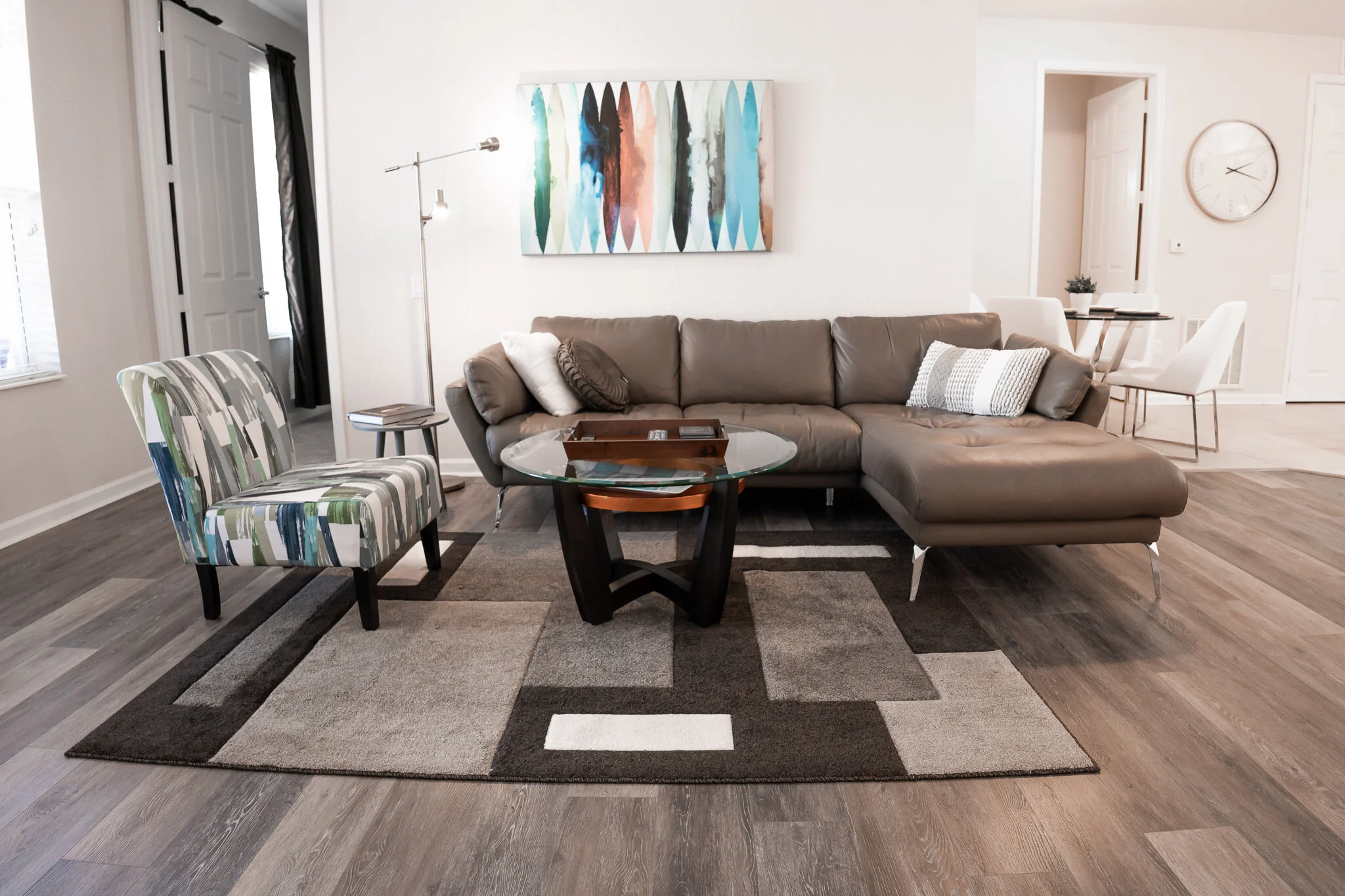 Living room with a brown sectional sofa, a striped armchair, a glass coffee table, and a multicolored abstract painting on the wall. There is a geometric area rug and hardwood flooring.