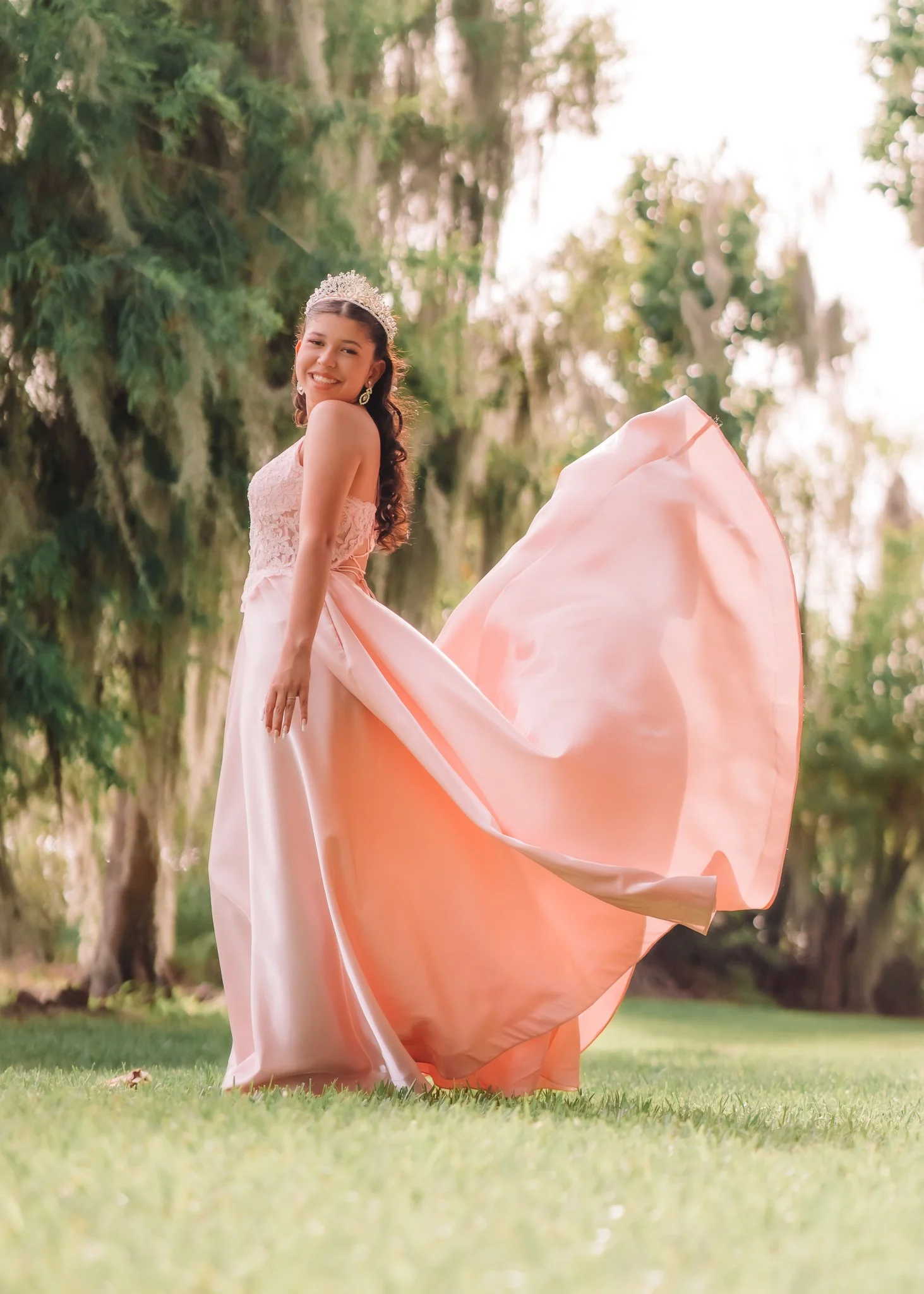 Young woman in a pink ball gown and tiara standing outdoors on grass, smiling