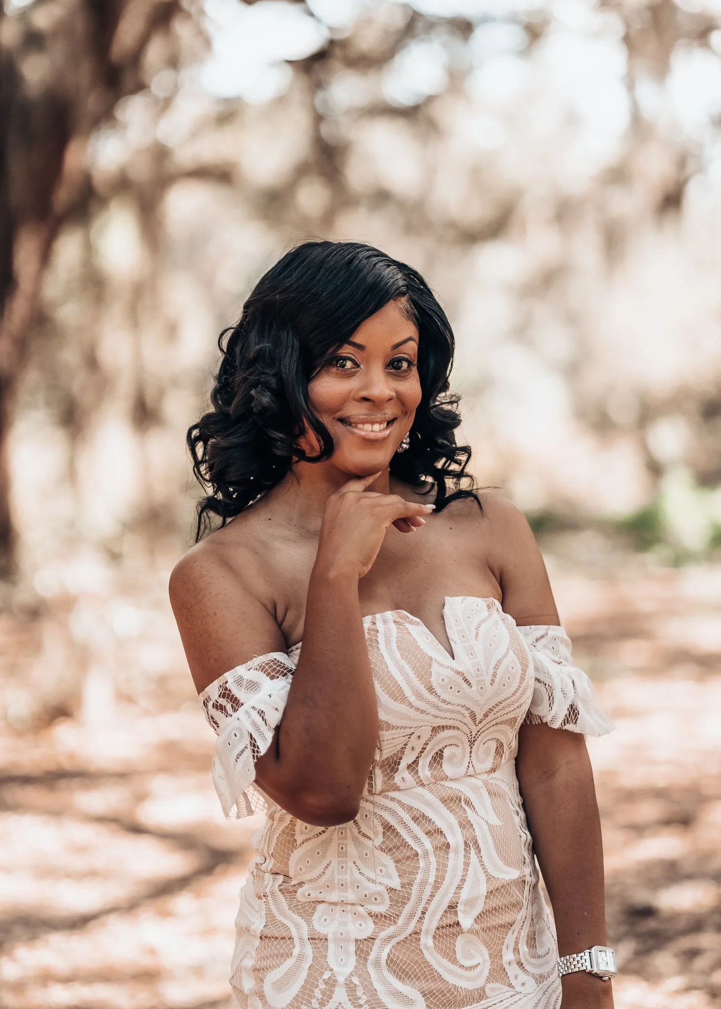 A woman in an off-the-shoulder white lace dress standing outdoors among blurred trees, smiling, with one hand resting near her chin.