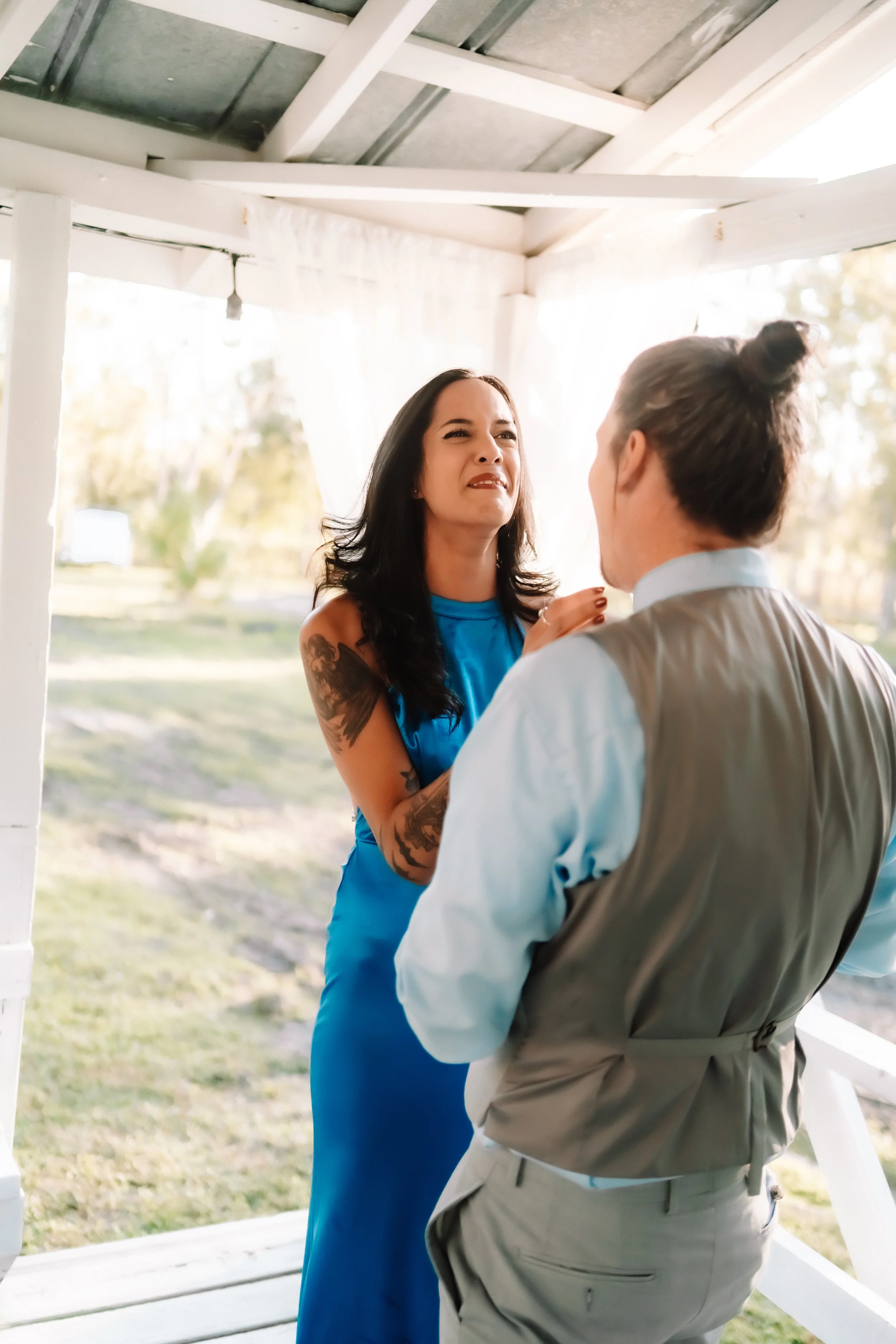 A woman with black hair, tattoos on her arm, wearing a blue dress, appears upset or angry at a man with light brown hair tied in a bun, wearing a gray vest and white shirt, on a porch with white railings and outside trees and grass in the background.