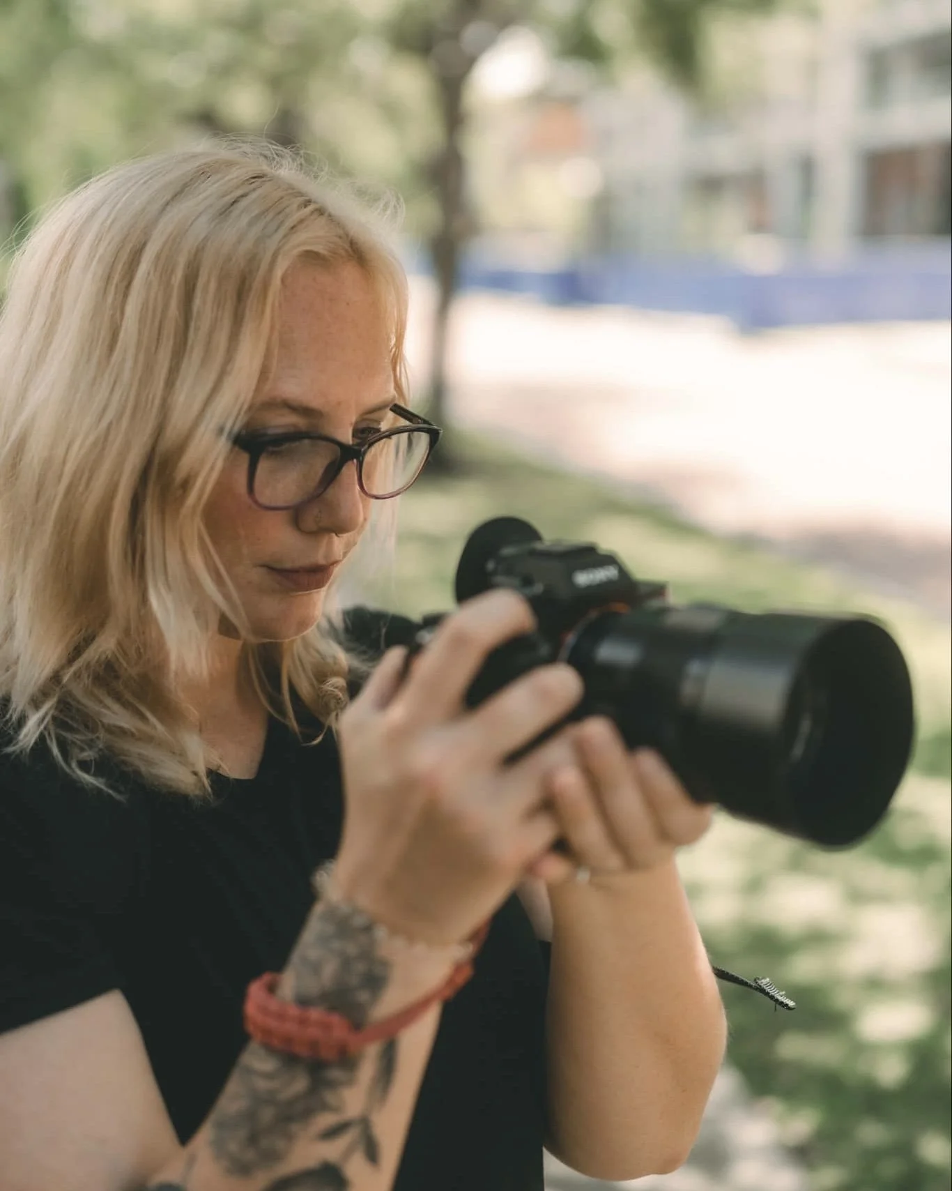 A woman with blonde hair and glasses looking at a camera outdoors in a park.