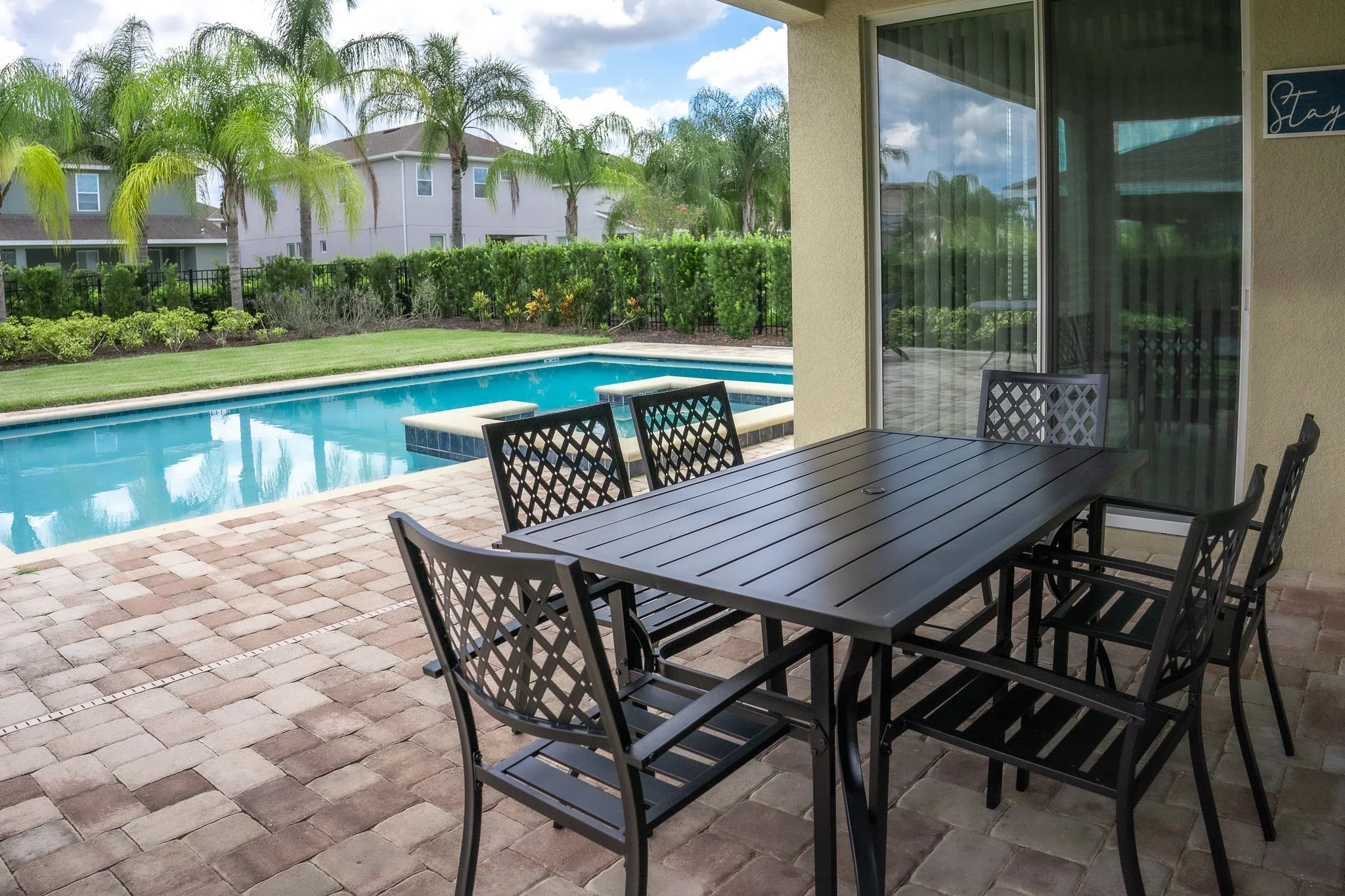 Patio with outdoor table and chairs overlooking a backyard swimming pool, lush green plants, and neighboring residential buildings.