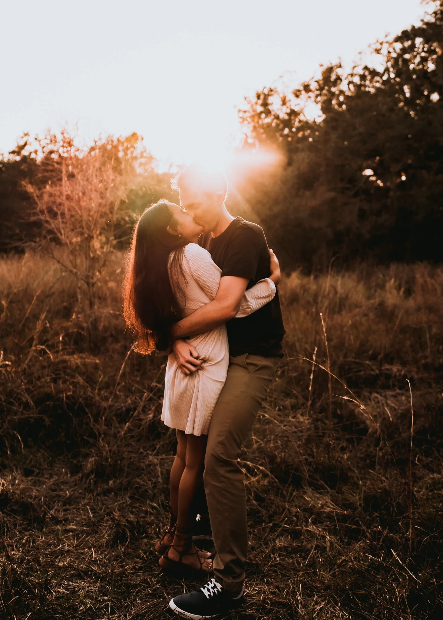 A couple embraces and kisses in an outdoor setting during sunset, with the sun shining behind them. The woman has long dark hair and wears a beige dress. The man has short hair and wears a black t-shirt, khaki pants, and black sneakers. They are stan