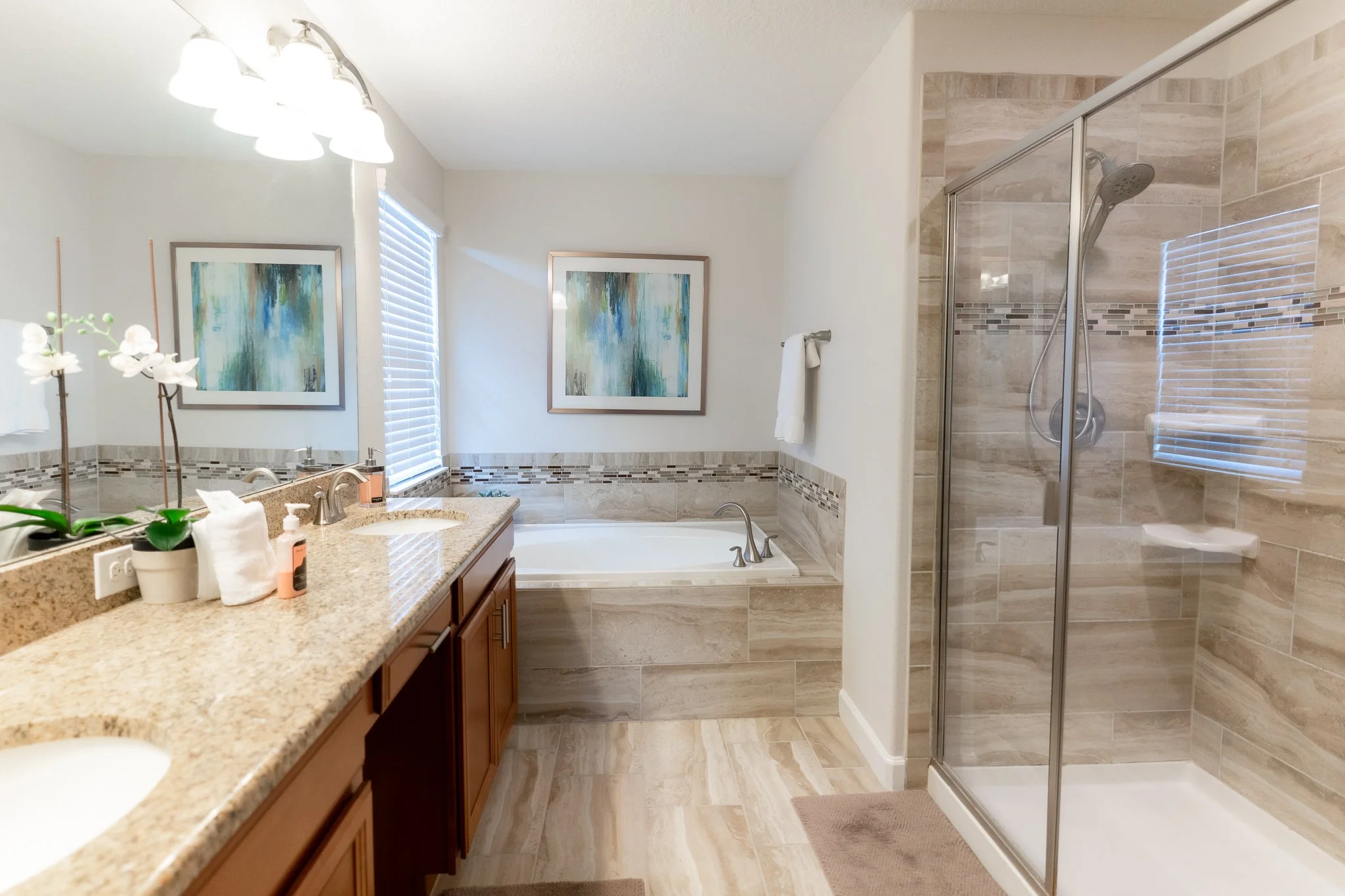 A modern bathroom with a granite countertop, wooden cabinets, a double sink, a large mirror, a bathtub with artwork above it, and a separate glass-enclosed shower with beige and brown tile walls.