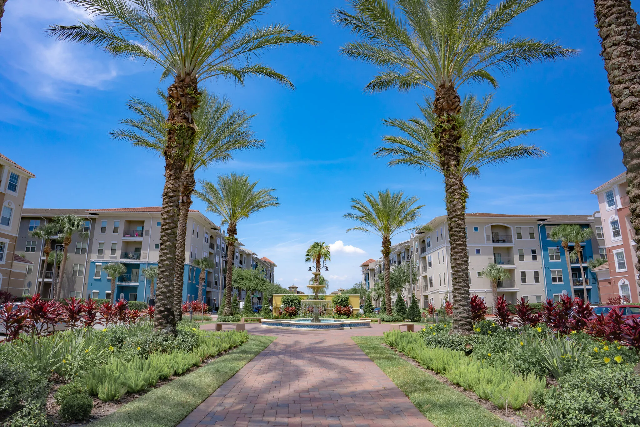 Colorful apartment complex courtyard with a brick pathway, lush landscaping, palm trees, and a multi-tiered fountain under a bright blue sky.