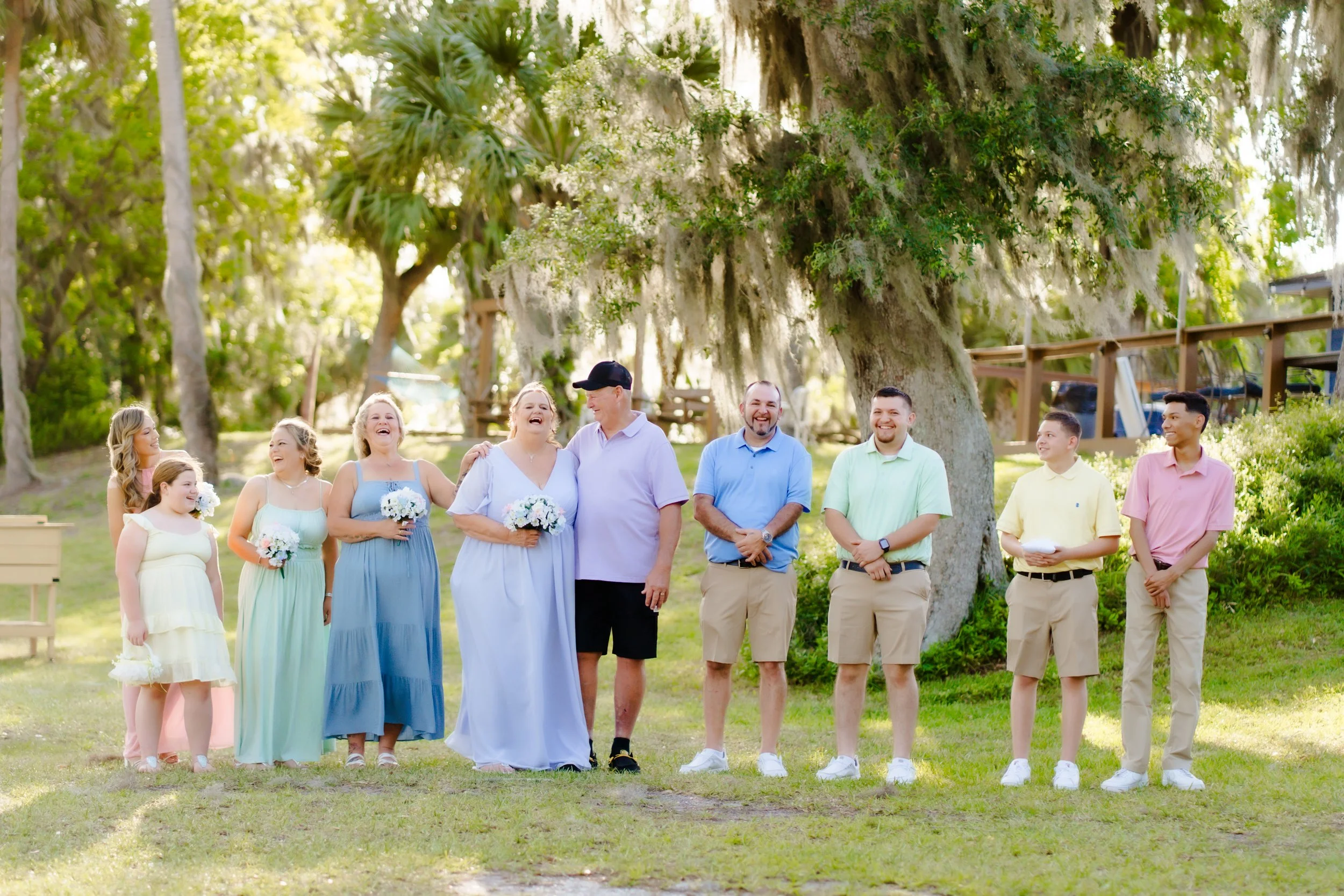 A group of people, including women in pastel dresses and men in casual shirts, standing outdoors under large trees during what appears to be a wedding or celebration. Some are holding small bouquets, and everyone is smiling and looking joyful.