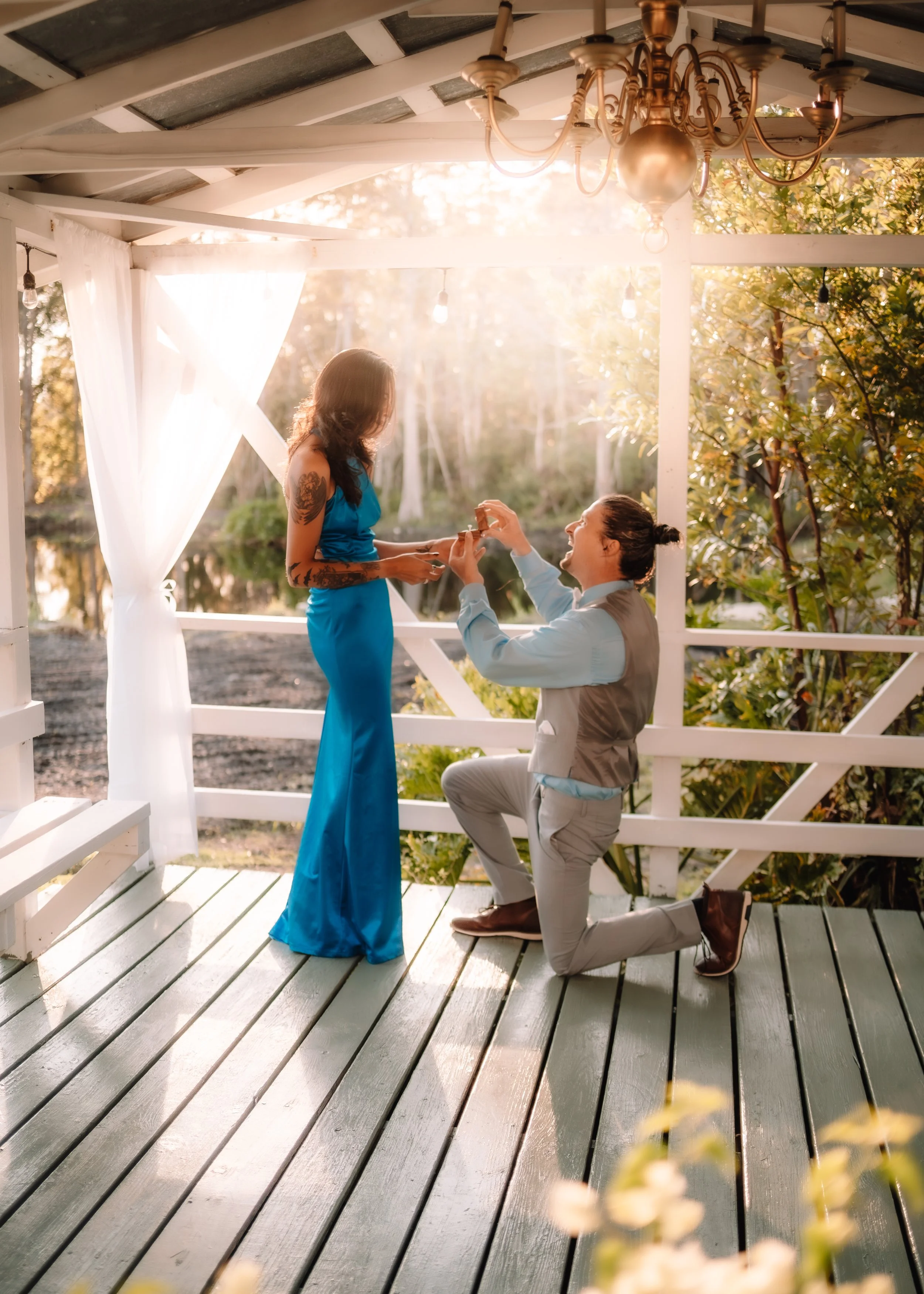 A man kneeling and proposing to a woman in an outdoor gazebo during sunset, with the woman wearing a blue dress and the man in a vest and tie, surrounded by trees and warm lighting.