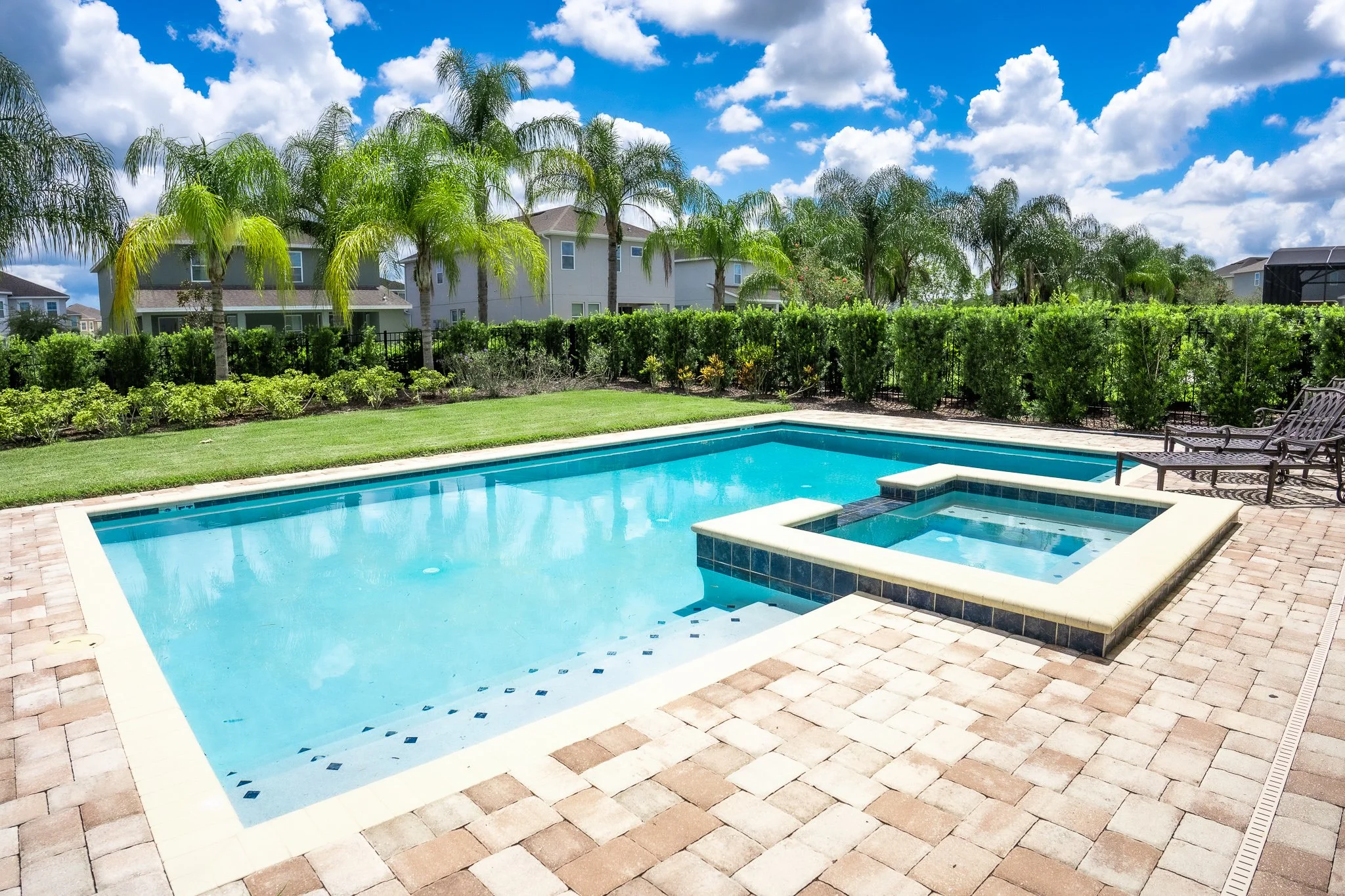 Residential backyard with a rectangular swimming pool and a hot tub, surrounded by tan brick paving, green lawn, palm trees, and a row of houses in the background under a blue sky with white clouds.