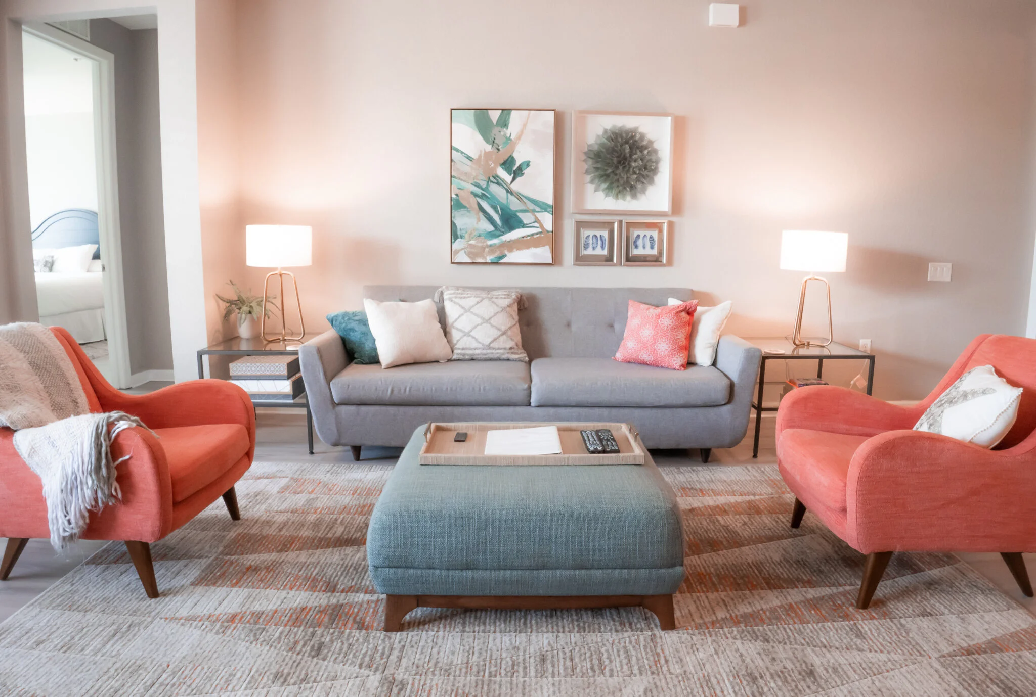 Living room with gray sofa, pink armchairs, and a green ottoman in the center. Two side tables with lamps, framed artwork, and decorative pillows. A bedroom is partially visible through an open door.