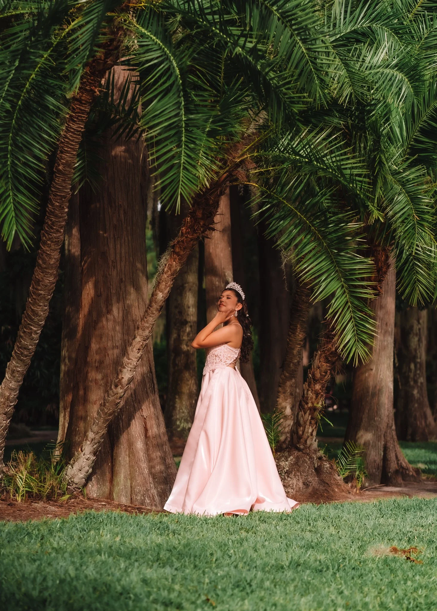 A young woman in a pink gown and tiara standing among large tropical trees in a forest setting.