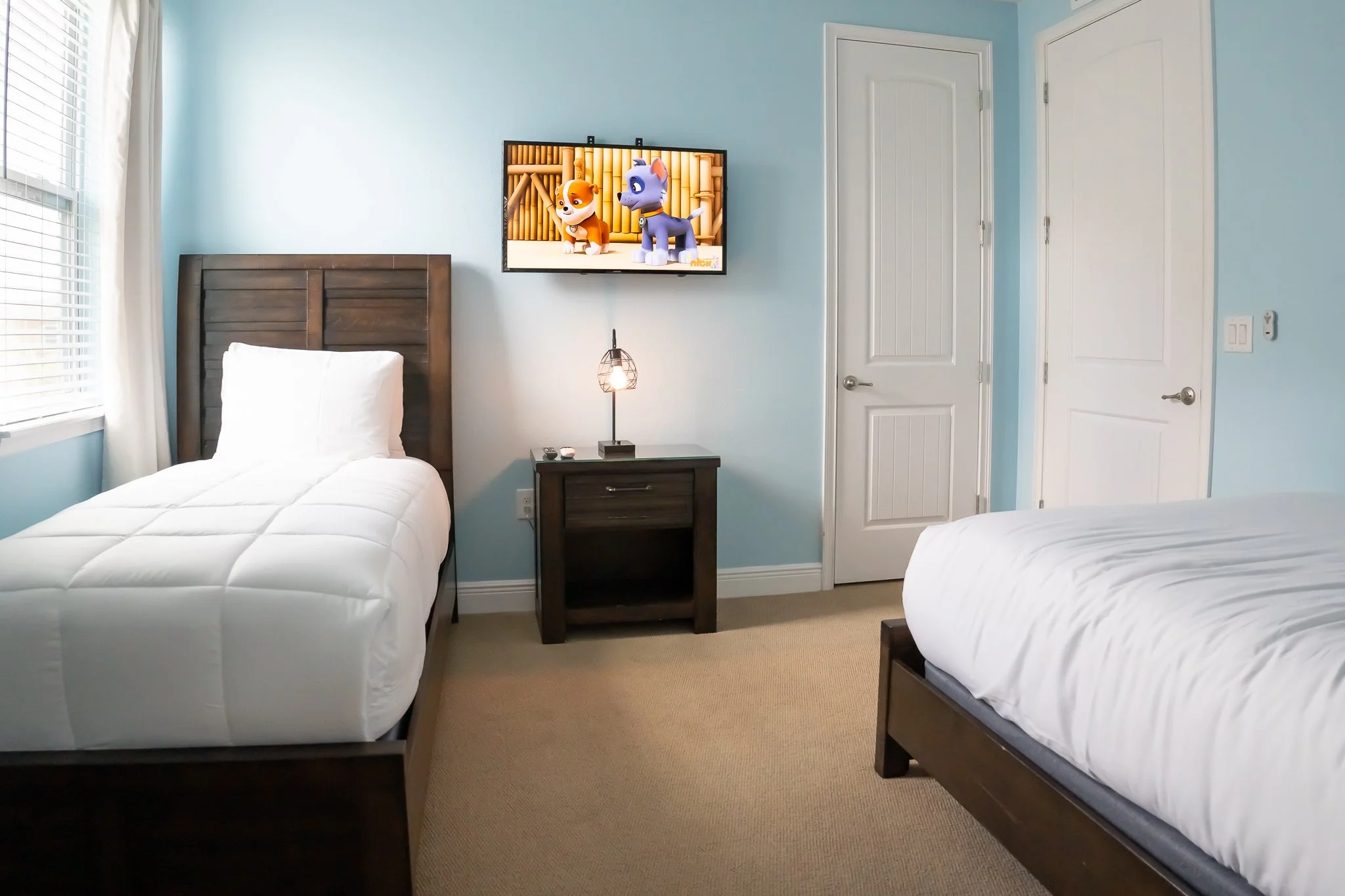 A bedroom with two beds with white bedding, light blue walls, and a window with white blinds. There is a dark wood nightstand between the beds with a small lamp and a TV mounted above it. The TV is showing animated characters. There are two white doo