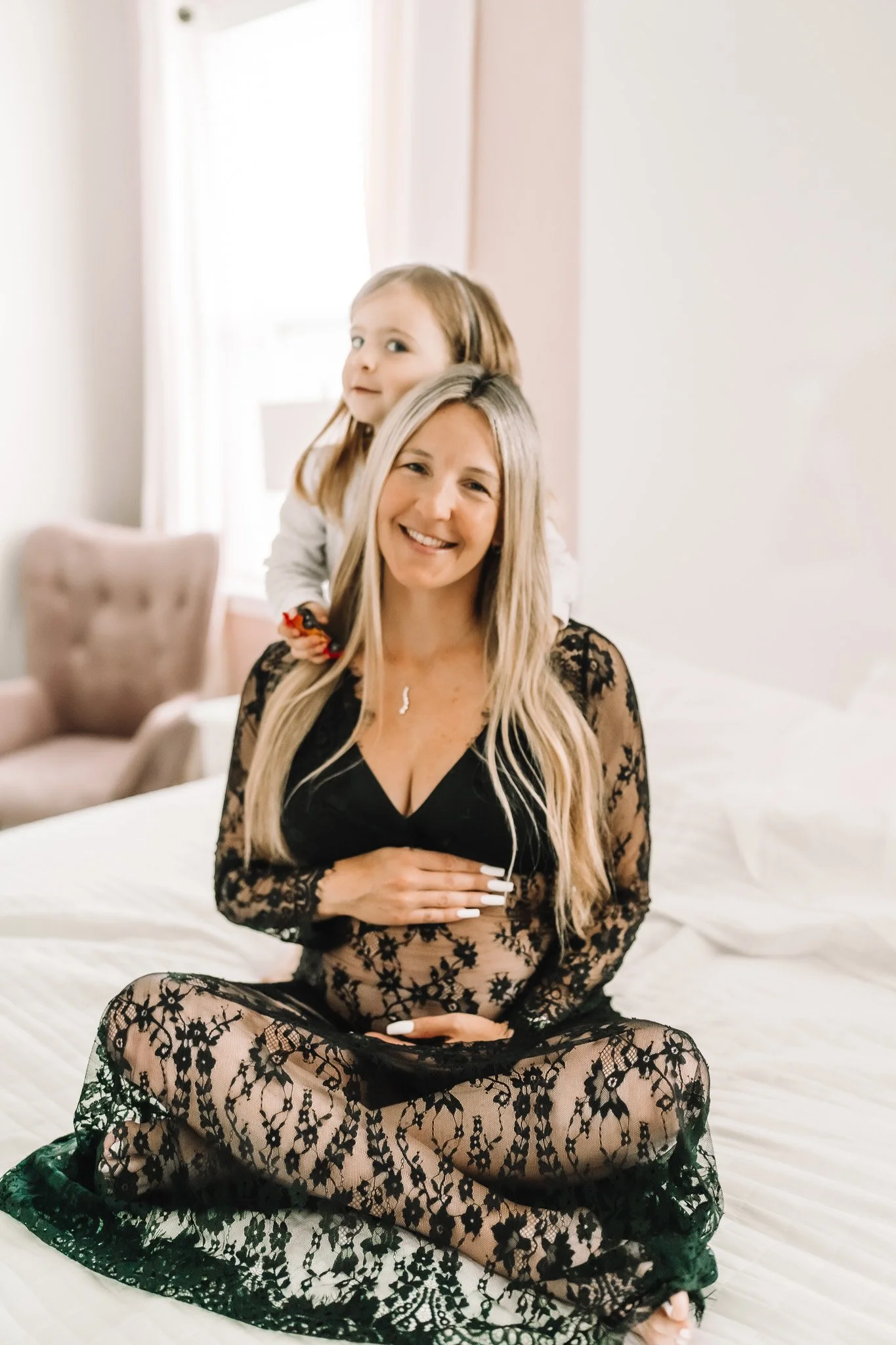 A woman in black floral lace lingerie sitting on a bed with a young girl on her shoulders in a bright, softly lit room.