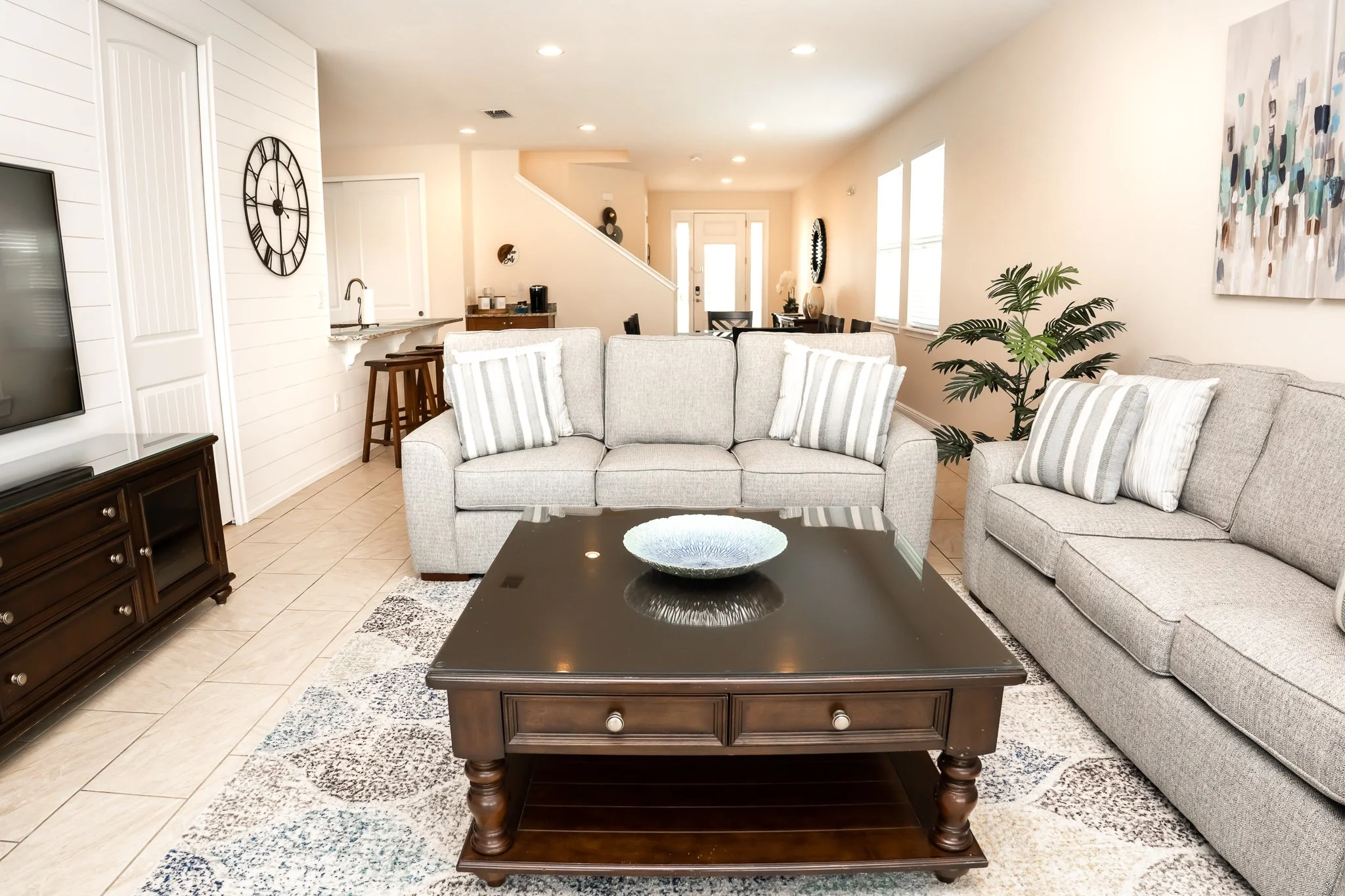 Living room with two light gray sofas, a dark wooden coffee table, a patterned area rug, a large wall clock, a potted plant, and abstract artwork on the wall.