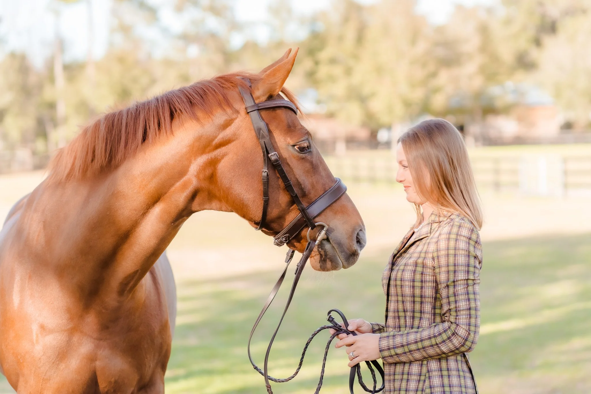 A woman in a plaid blazer standing outside with a brown horse, holding the horse's reins, in a green field with trees in the background.