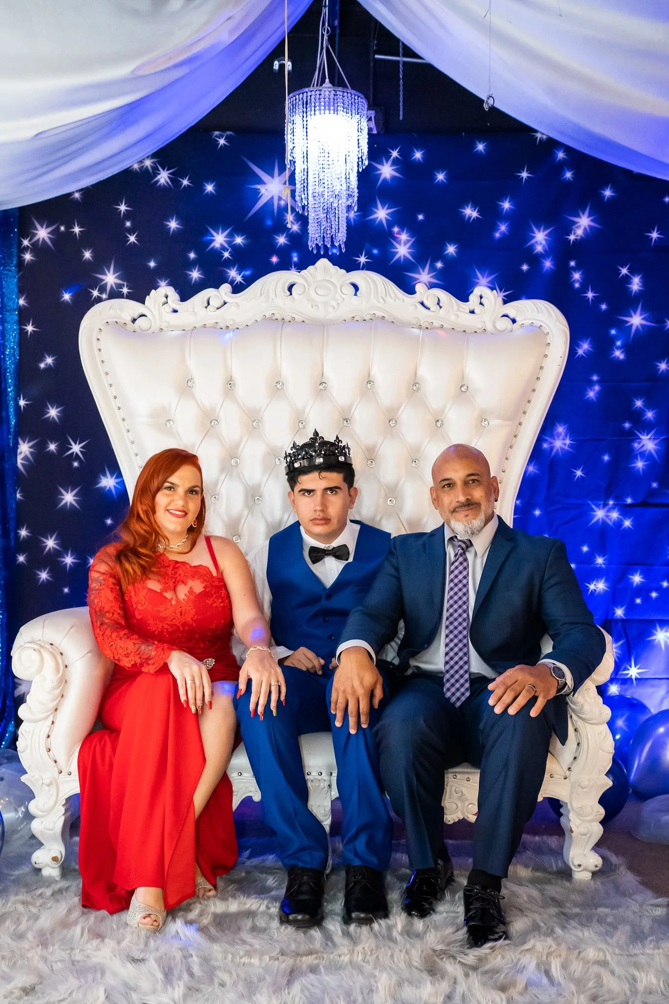 Three people sitting on a white ornate throne-like sofa at a festive event. The woman on the left has long red hair and is wearing a red lace dress. The man in the middle has dark hair, a serious expression, and is wearing a blue tuxedo with a black 