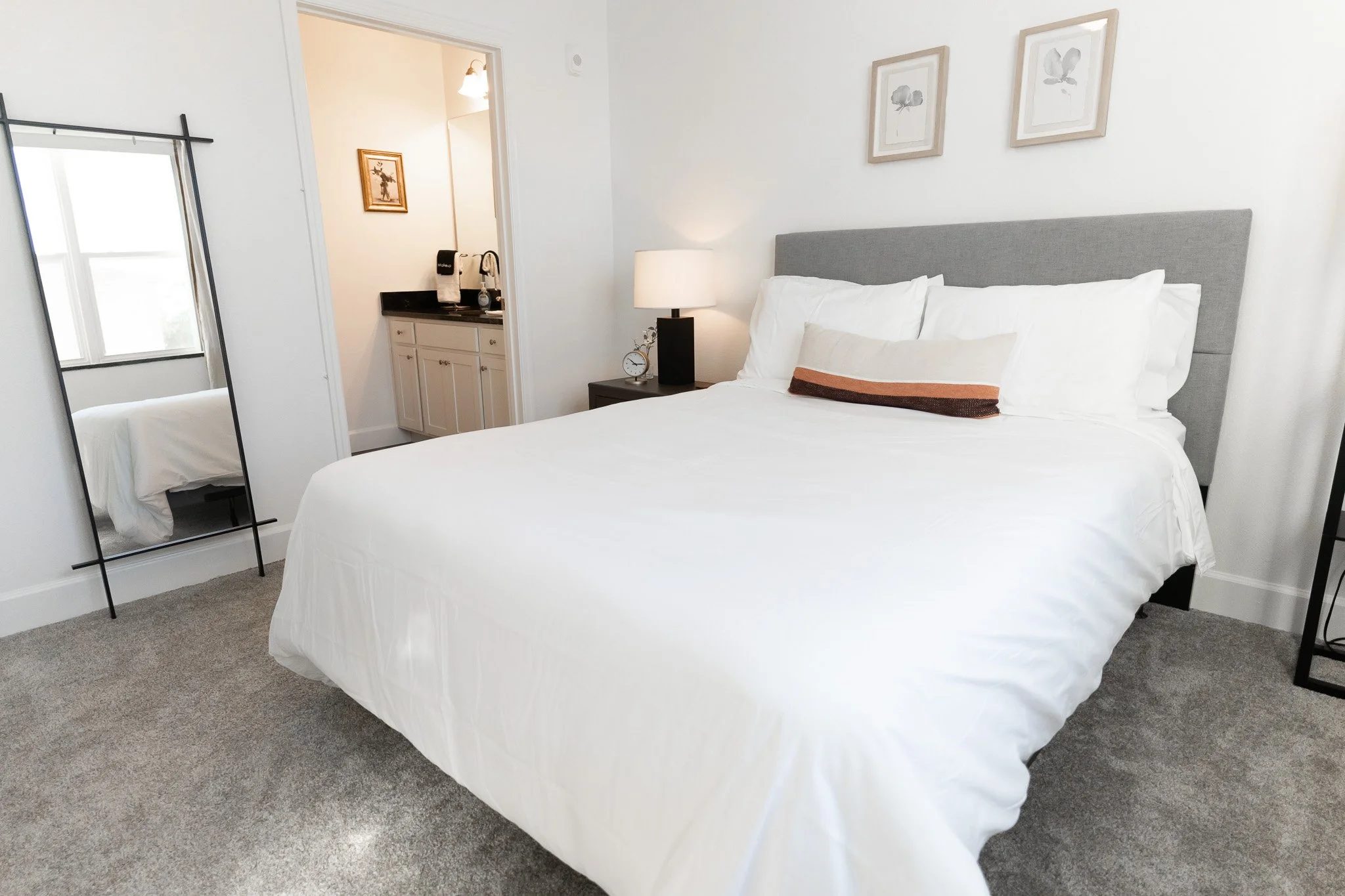 A neatly made bed with white bedding and pillows, gray headboard, bedside table with a lamp and an alarm clock, framed art on the wall, a full-length mirror, and a glimpse of a bathroom with a sink and a coffee maker.