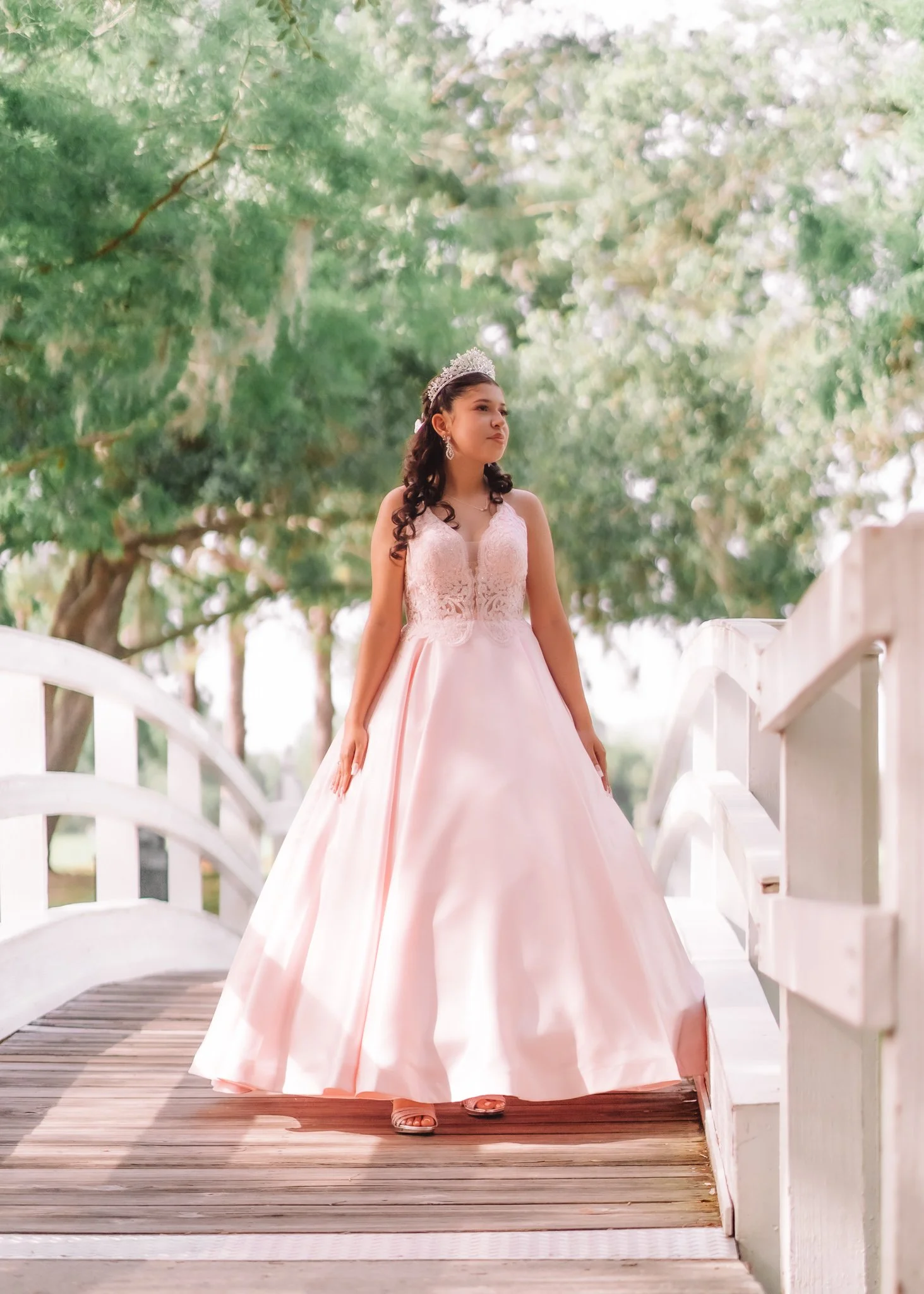 A girl wearing a pink princess-style dress, tiara, and earrings stands on a small arched wooden bridge surrounded by lush green trees.