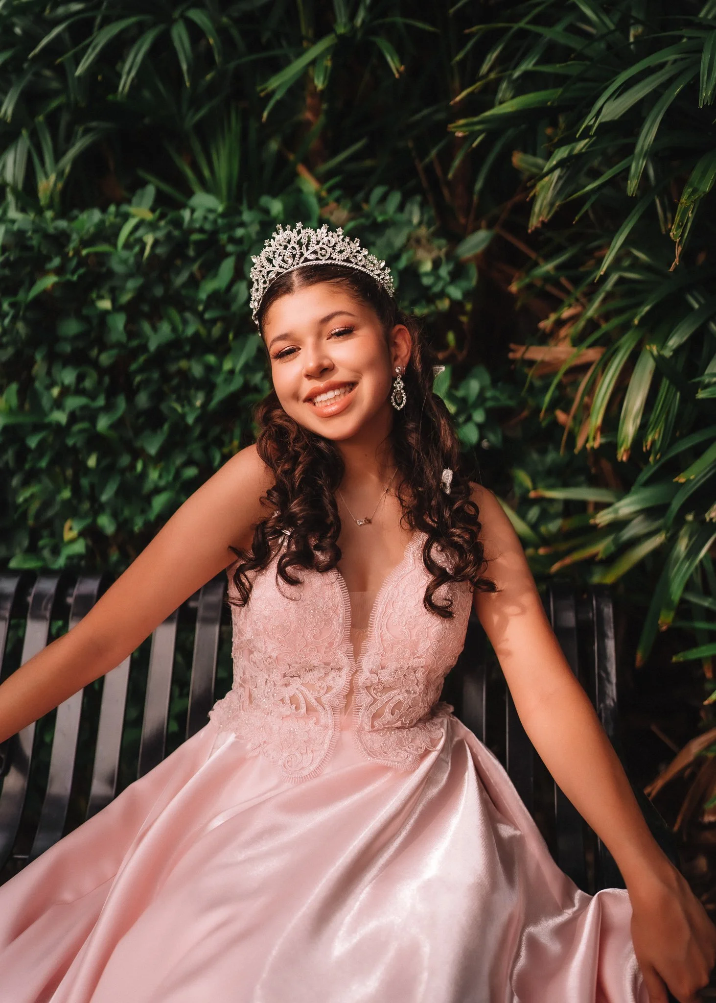 Young woman wearing a pink dress, tiara, and earrings, sitting on a black bench in front of green foliage, smiling at the camera.