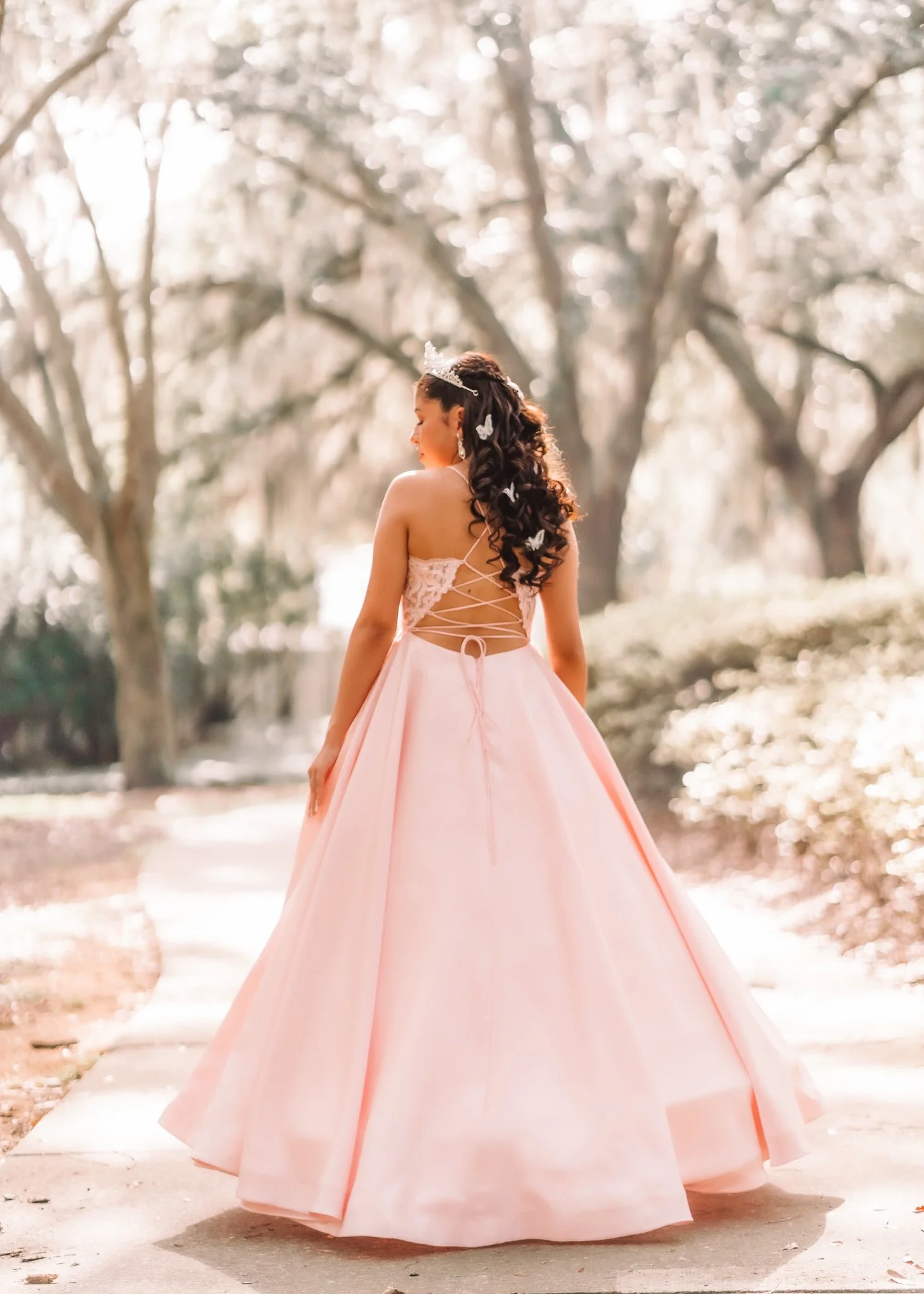 A woman in a pink ball gown with lace and crisscross back detail, standing outdoors on a sidewalk, with trees and sunlight in the background.