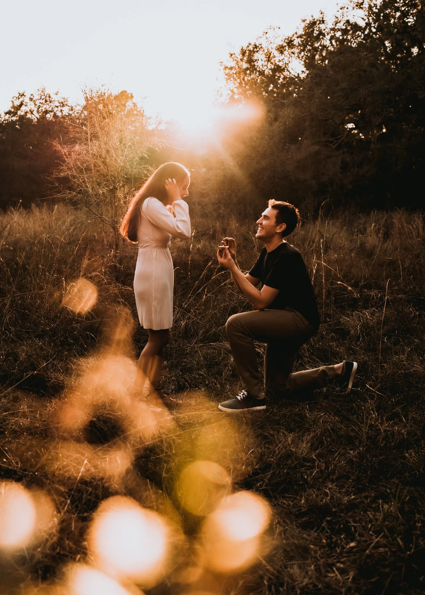 A man proposing marriage to a woman in a grassy outdoor setting during sunset.