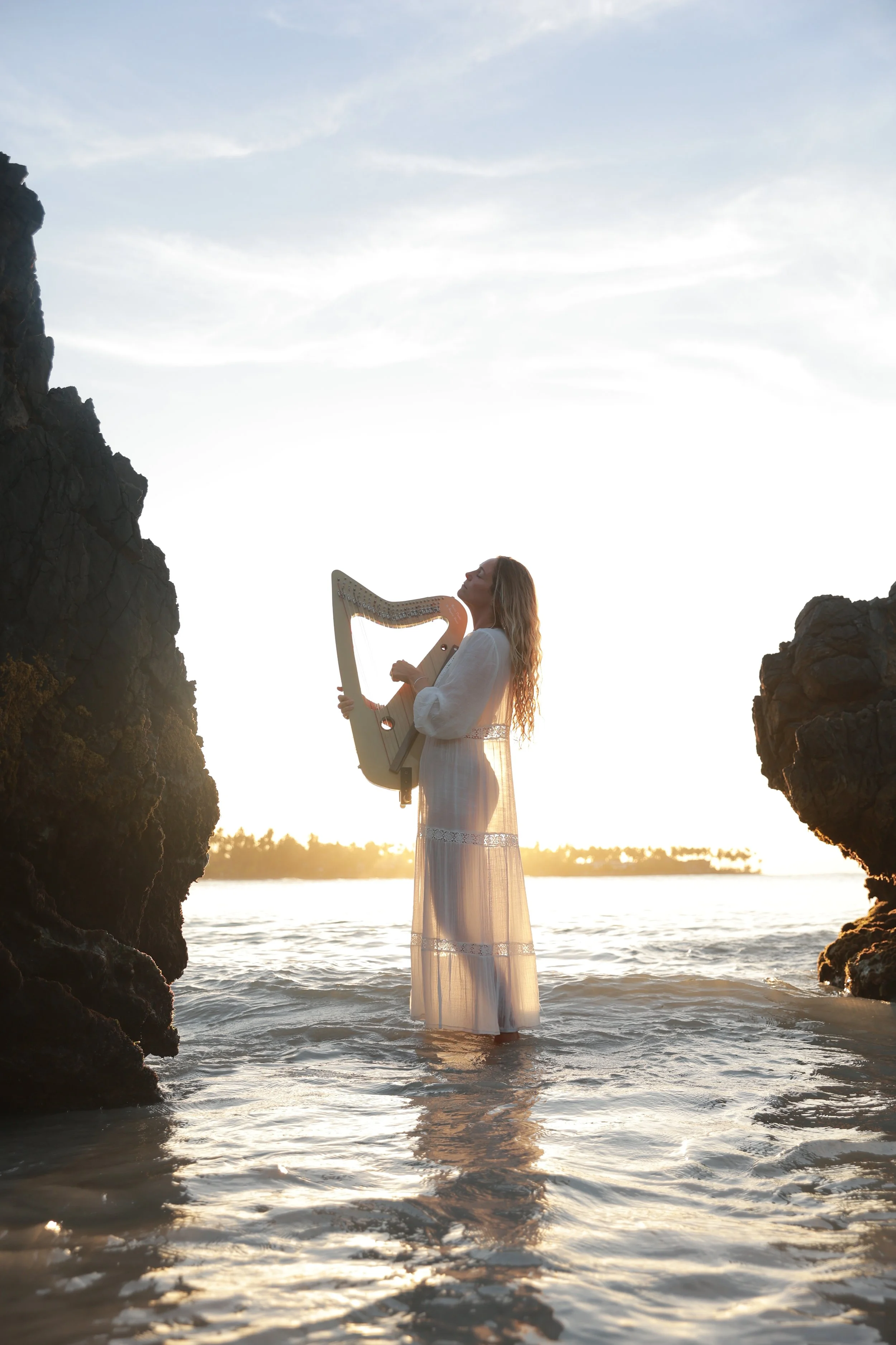 Lifestyle model playing harp in the ocean at sunset between coastal rocks, artistic tropical lifestyle scene.