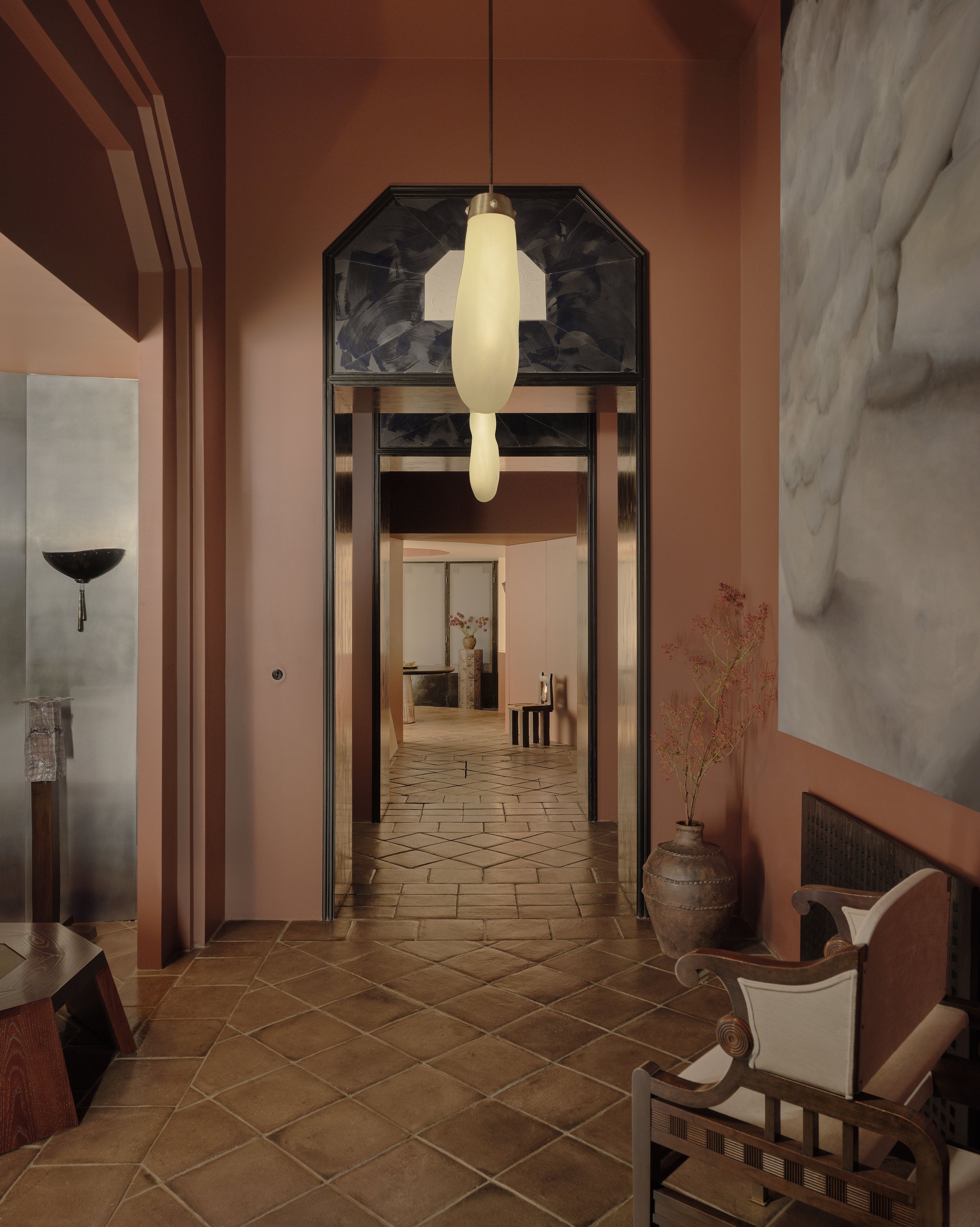 Interior view of a hallway with terracotta tile flooring, pink walls, a tall vase with a plant, a chair, and a large painting, leading to a room with another vase and flowers.