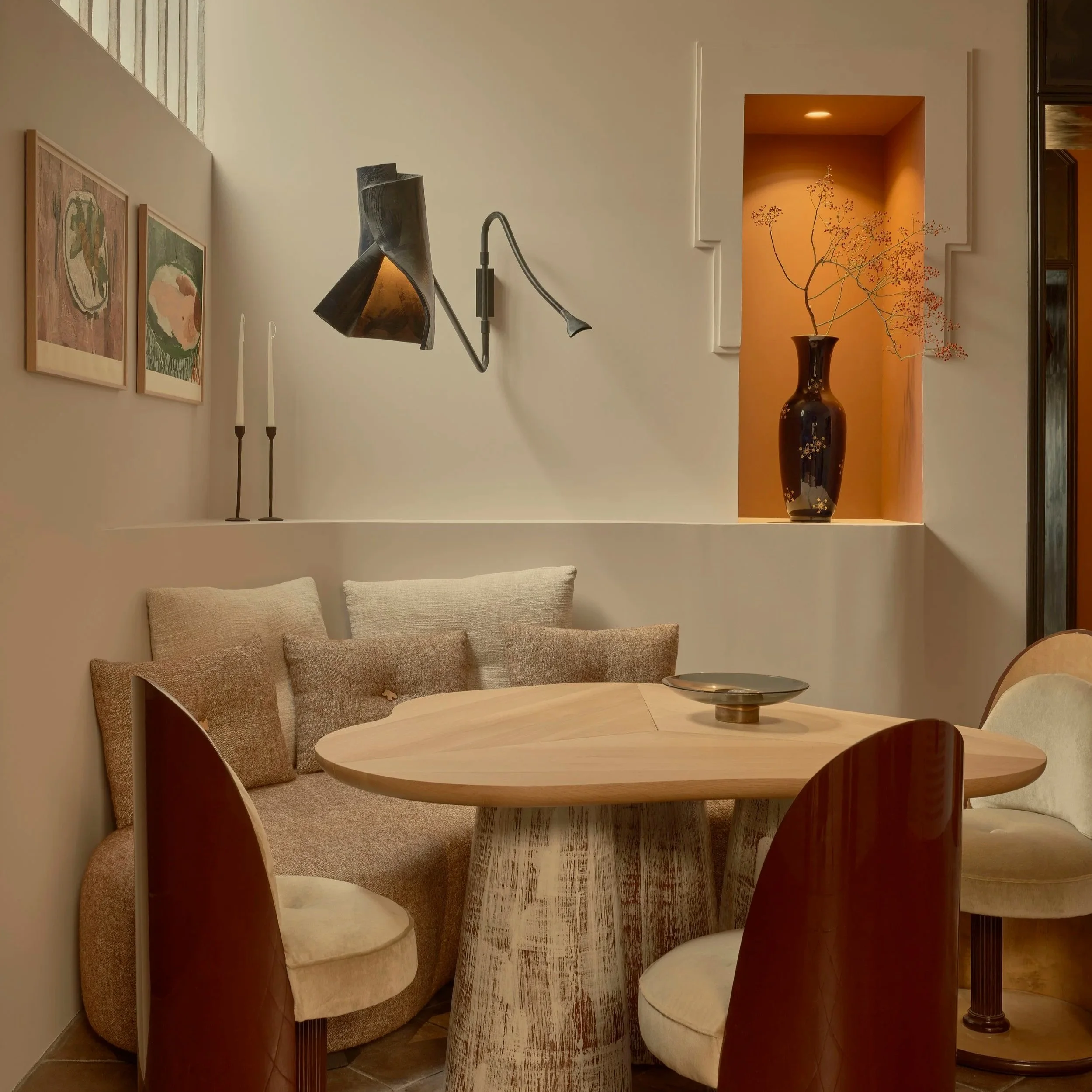 A cozy dining corner with a round wooden table, surrounding chairs, a cushioned bench with multiple pillows, wall-mounted lighting, framed artwork, and a built-in shelf with a decorative vase and branch.