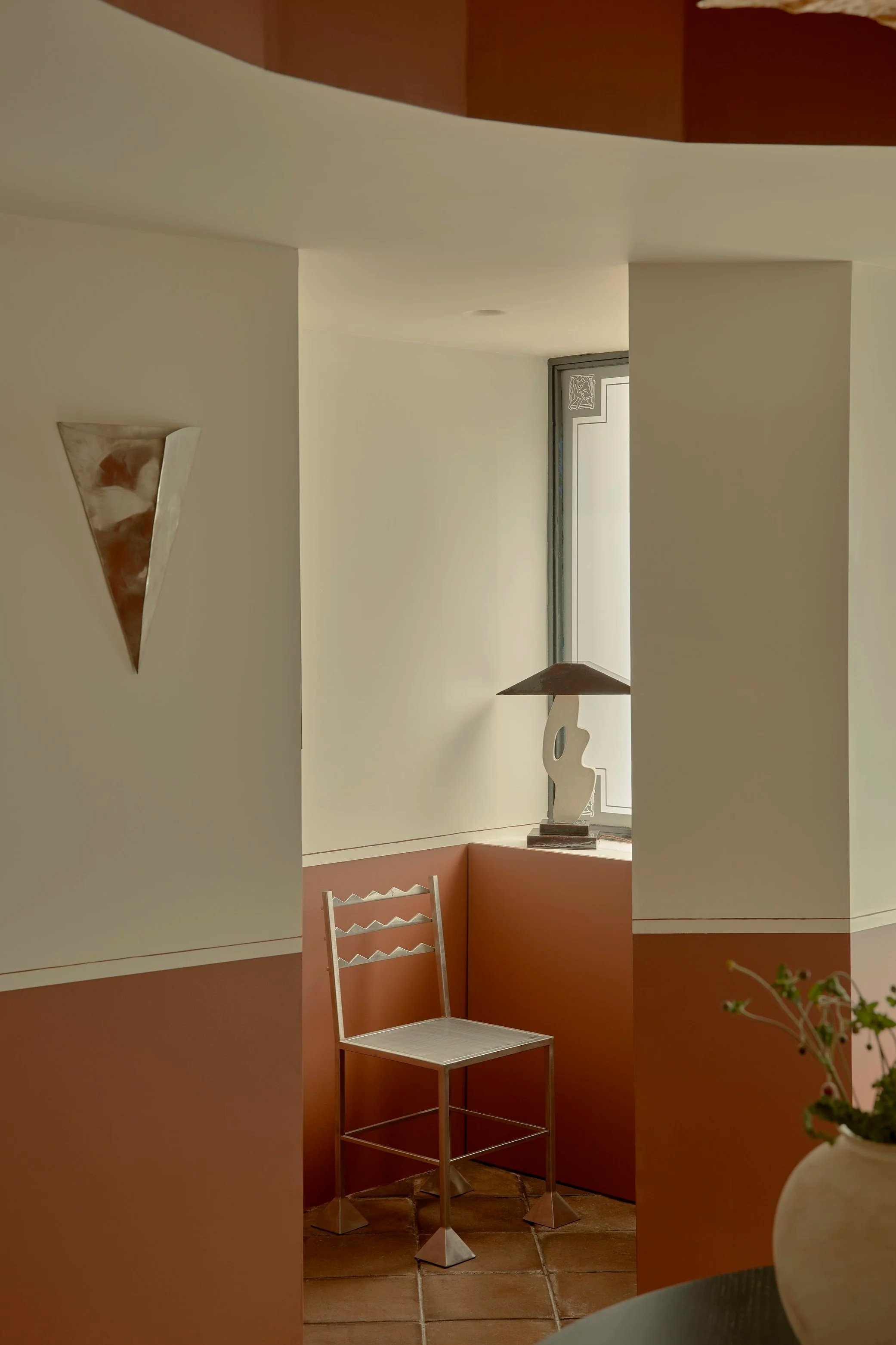 Interior corner of a room with a chair, a decorative sculpture on the window ledge, a wall-mounted mirror, and a potted plant in the foreground.
