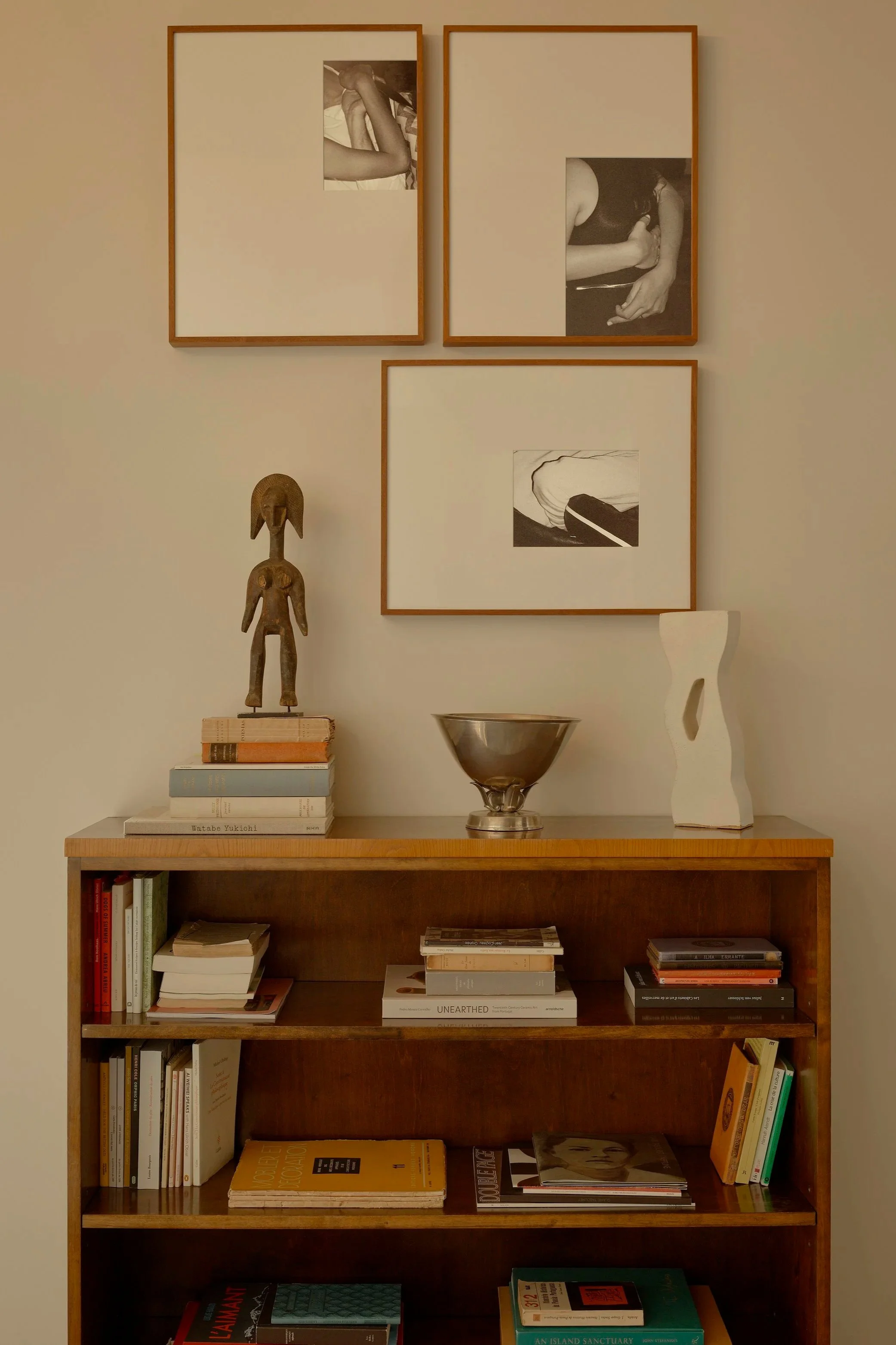 A wooden bookshelf with books and art objects on top. Three framed black-and-white photos of art and human body parts are hung on the wall above. Art sculptures and a bowl are placed on the top of the bookshelf.