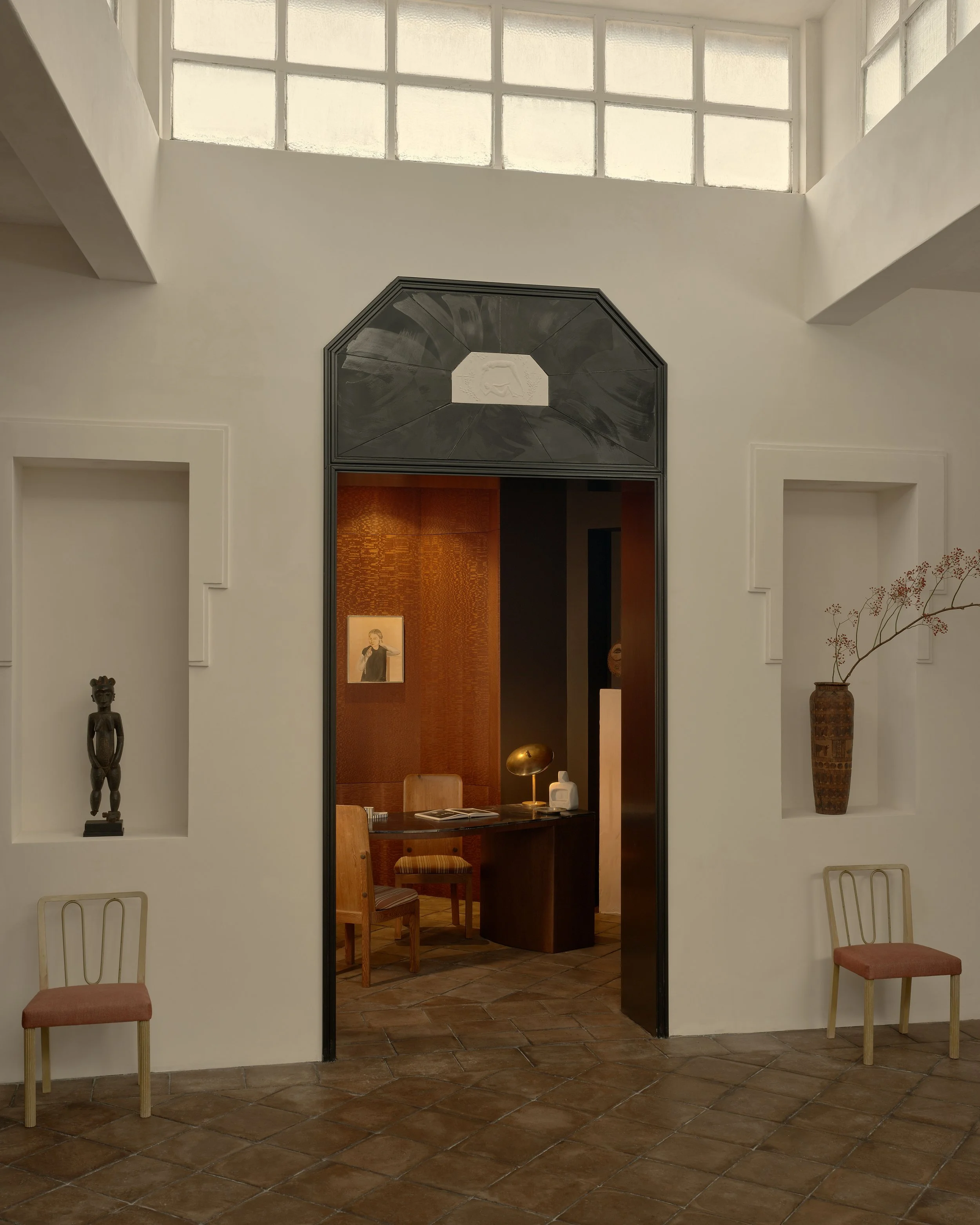 Interior of a room with white walls, decorative niches with art and vases, brick floor, and a glass block window at the top. Visible through a doorway is a cozy office space with wooden furniture, a desk, chairs, and warm lighting.