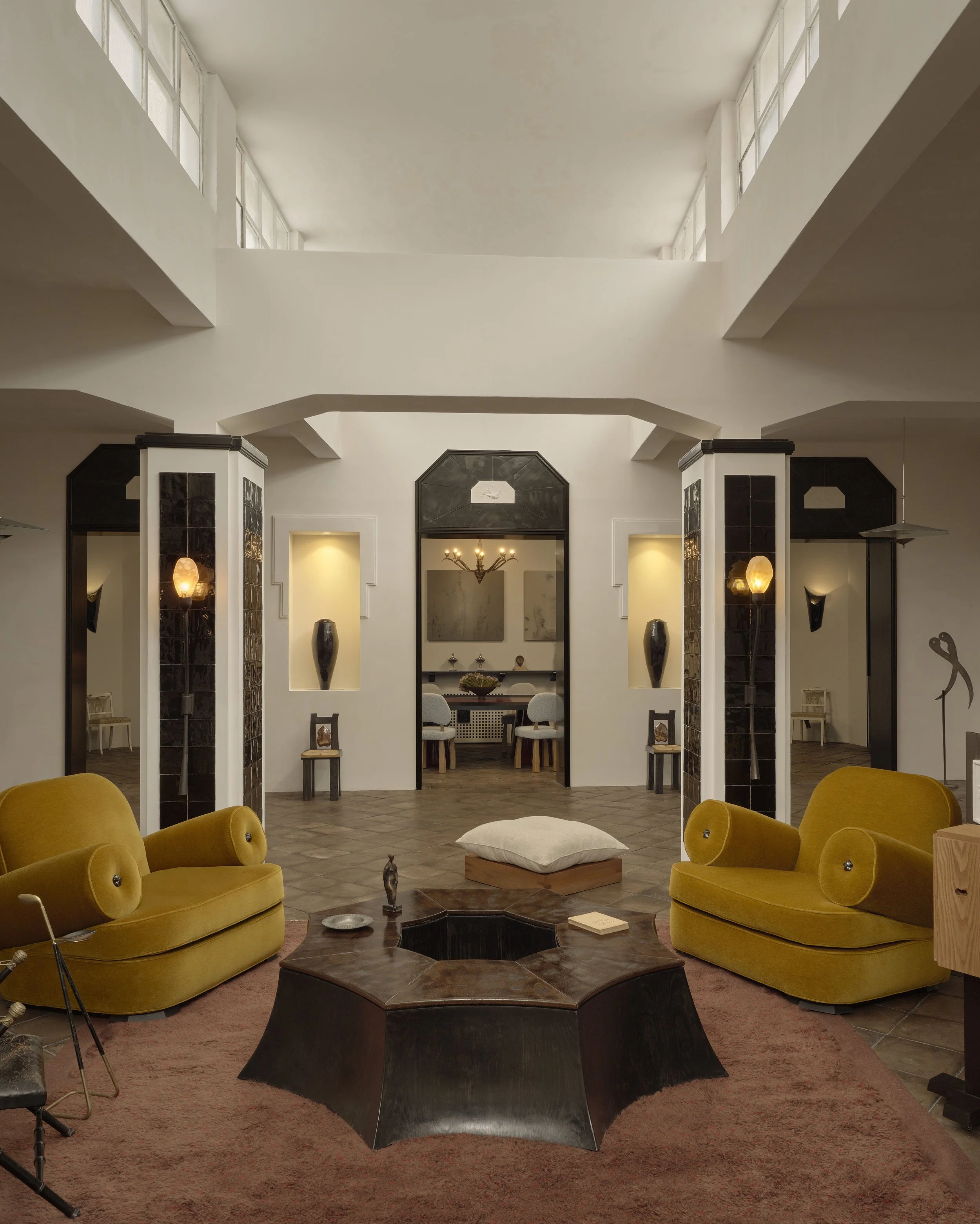 Living room with yellow sofas and a dark wooden coffee table with an octagonal hole, surrounded by a pinkish rug. In the background, a dining area with chairs and a chandelier, framed by black and white architectural features.