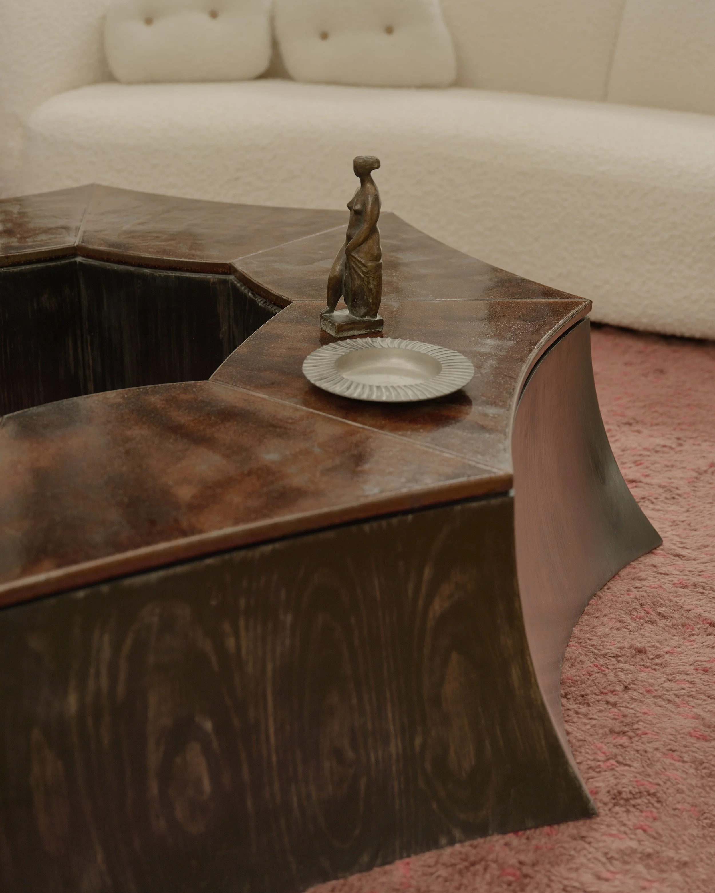 A corner of a wooden table with a small rustic sculpture of a woman and a silver plate on top, with a light-colored couch in the background.