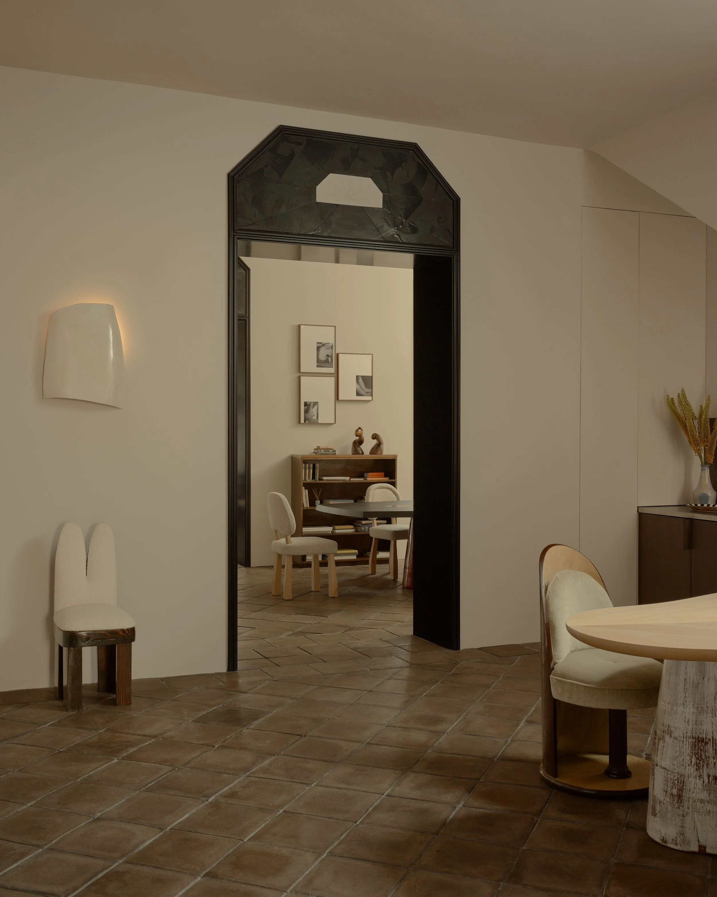 View of a cozy interior room with a tiled floor, modern wall sconce, and a black arched doorway leading to a room with framed artwork, a wooden bookshelf, and conversation chairs.