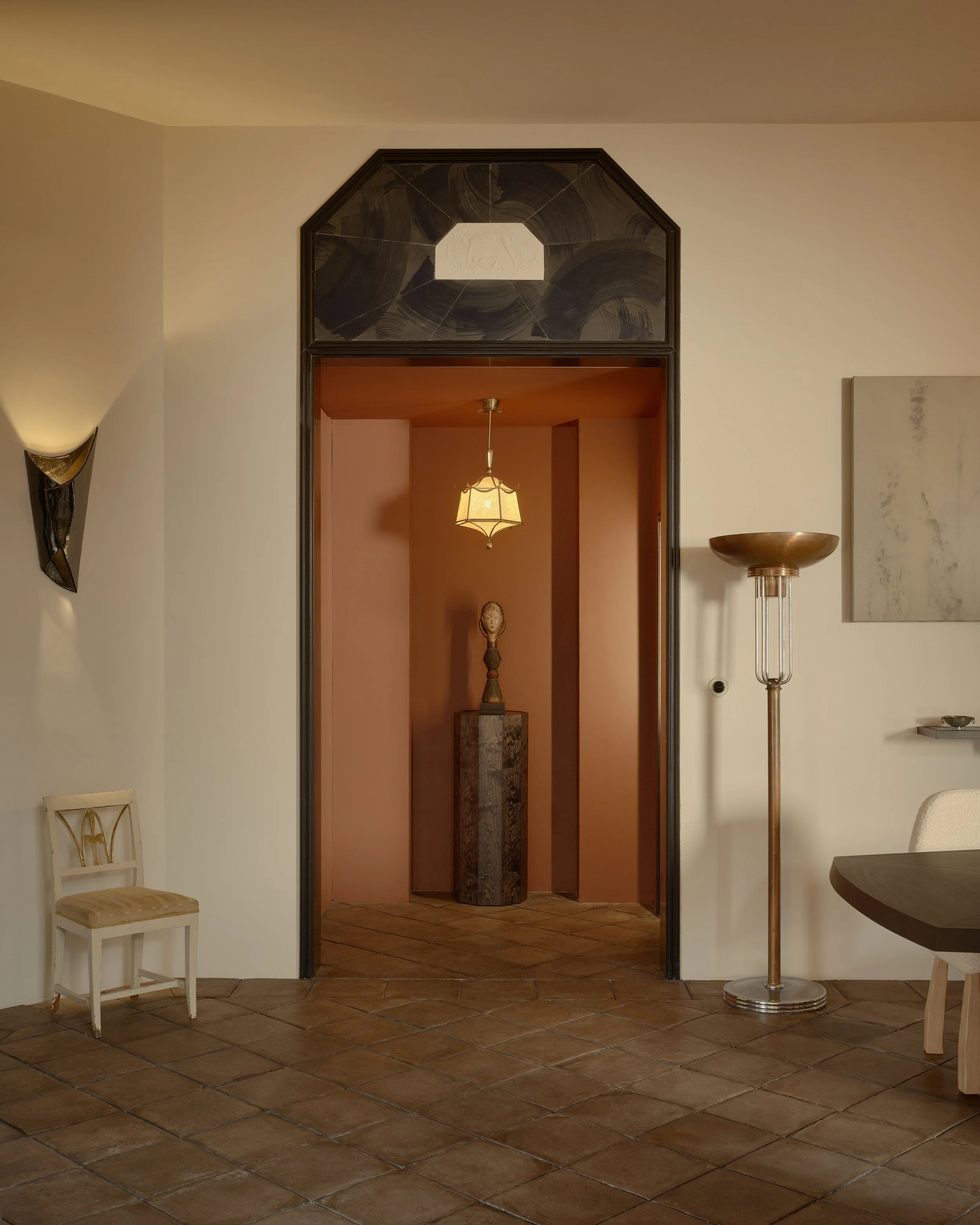 Interior view of a room with a doorway leading to a smaller space featuring a pedestal with a sculpture, with warm lighting and decorative furniture and art.