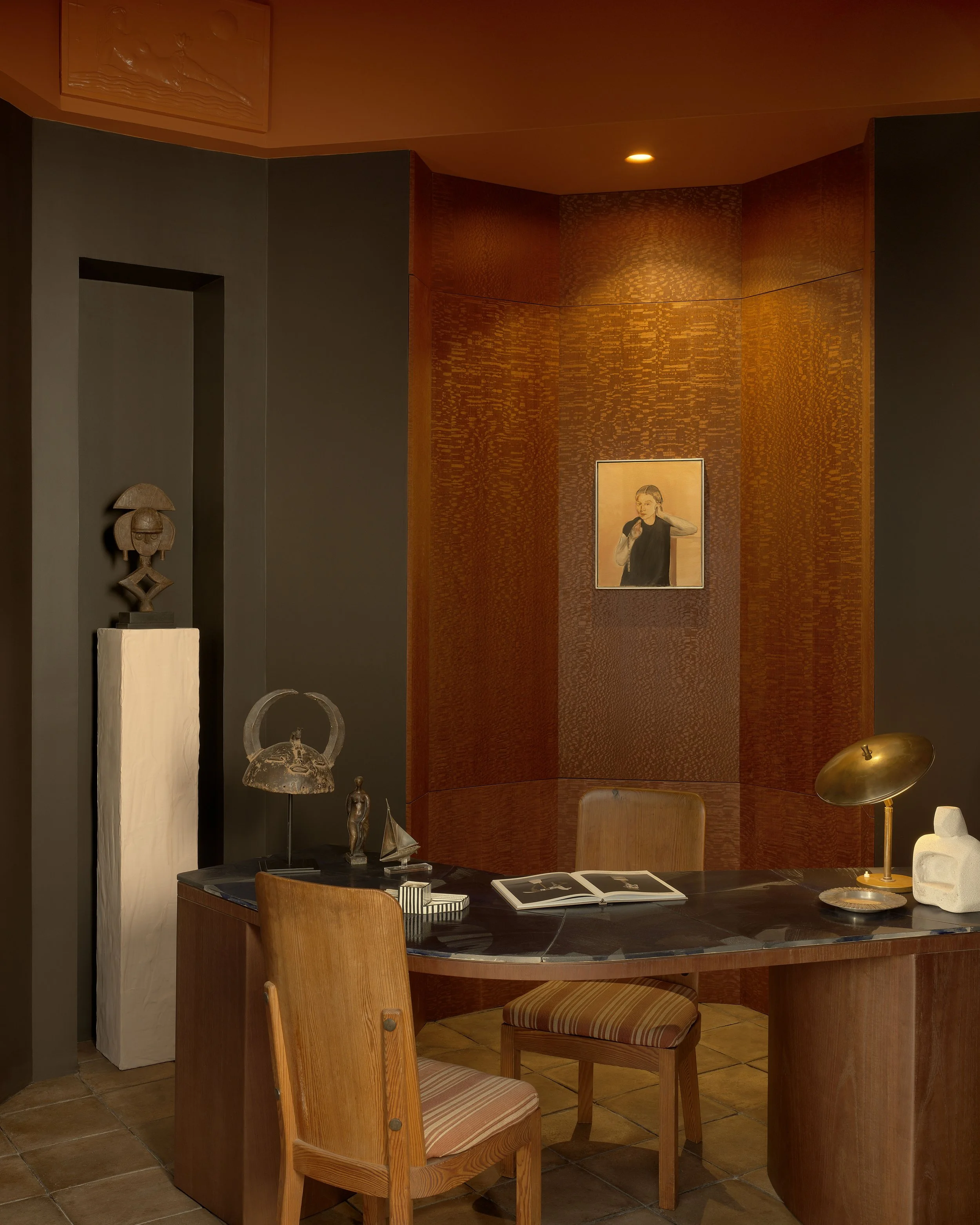 A cozy corner of a home office or study with a wooden desk, two wooden chairs with striped cushions, a black marble top on the desk, decorative sculptures, an open book, a gold desk lamp, a white abstract sculpture, and a framed portrait of a woman h