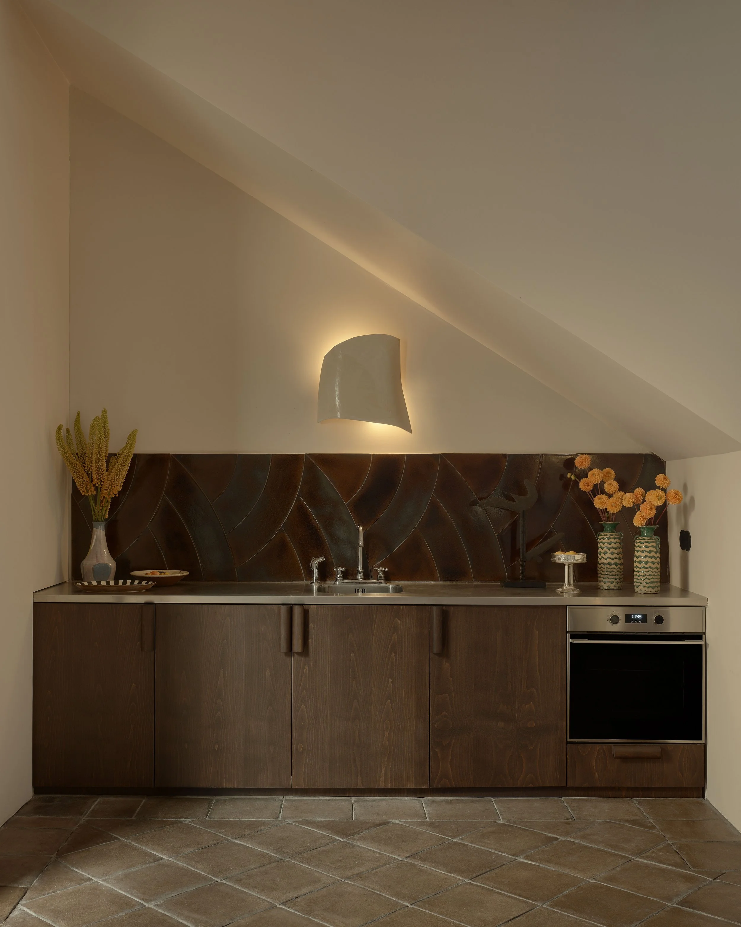 A minimalist kitchen with wooden cabinets, a small sink, a ceramic stove, and decorative vases with flowers on a countertop, against a sloped ceiling with a wall-mounted light fixture.
