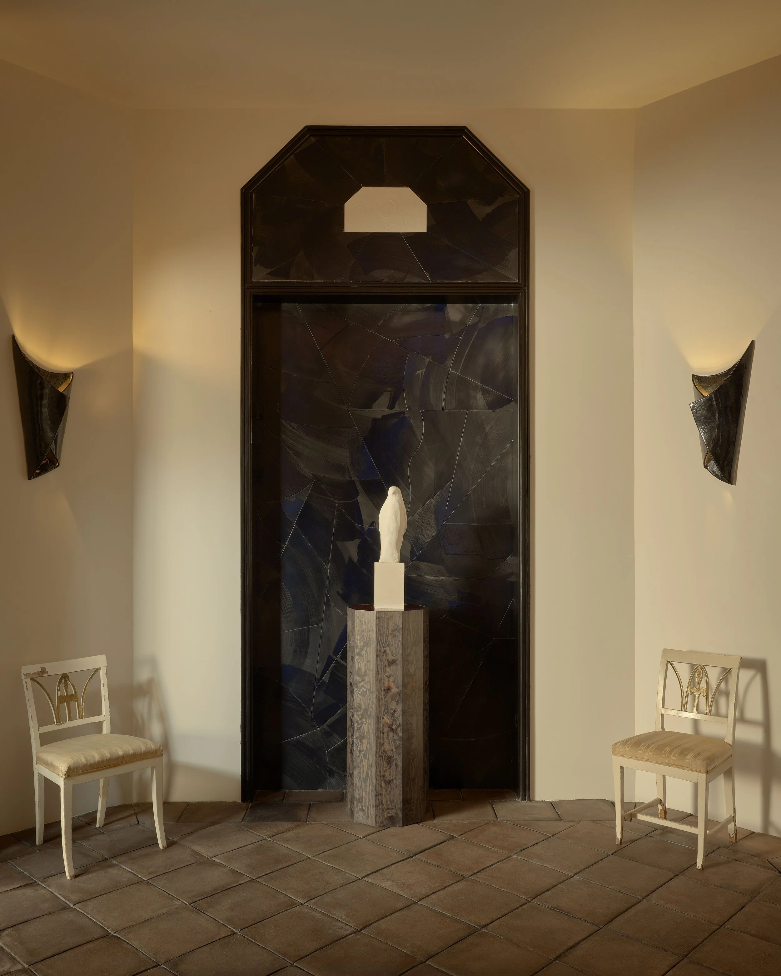 A minimalist art display in a room with beige walls and brown tiled floor. There is a black wall with a geometric pattern, two side sconces casting warm light, two white chairs with cushioned seats, and a central pedestal with a white sculpture of a 